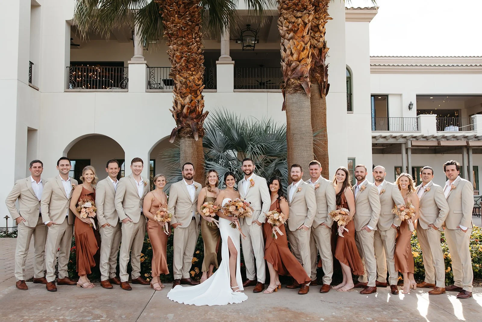 Full wedding party posing together beneath palm trees at La Quinta Resort with coordinated neutral attire and autumn-inspired florals. 