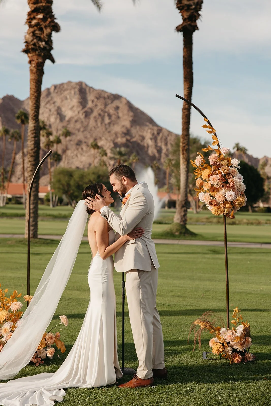  Bride and groom sharing their first kiss beneath minimalist floral installations with palm trees and mountain views at La Quinta Resort. 