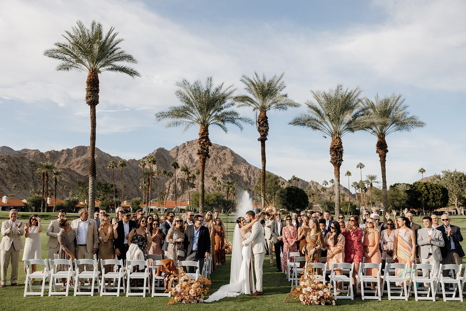  Wide view of the La Quinta Resort wedding ceremony as guests stand and cheer beneath palm trees and mountain scenery. 