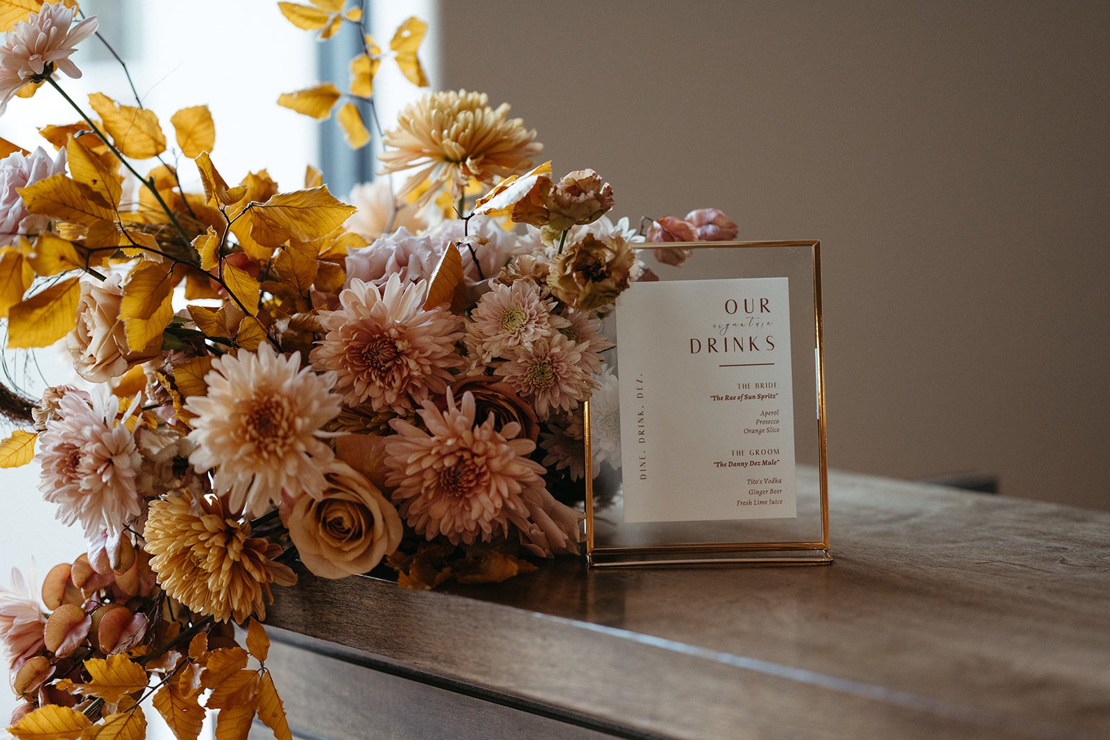  Seasonal floral arrangement in blush, peach, and warm golden tones styled beside a framed signature drinks sign at a La Quinta Resort wedding reception. 