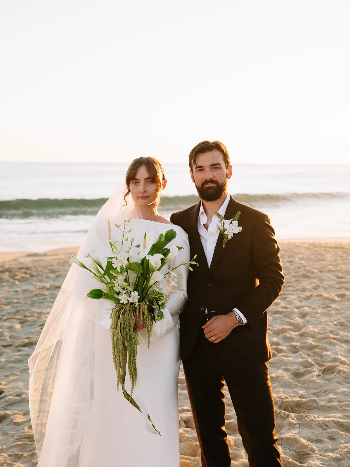 Bride and groom standing on the beach at sunset, the bride holding a modern white and green bouquet with calla lilies, orchids, and amaranthus