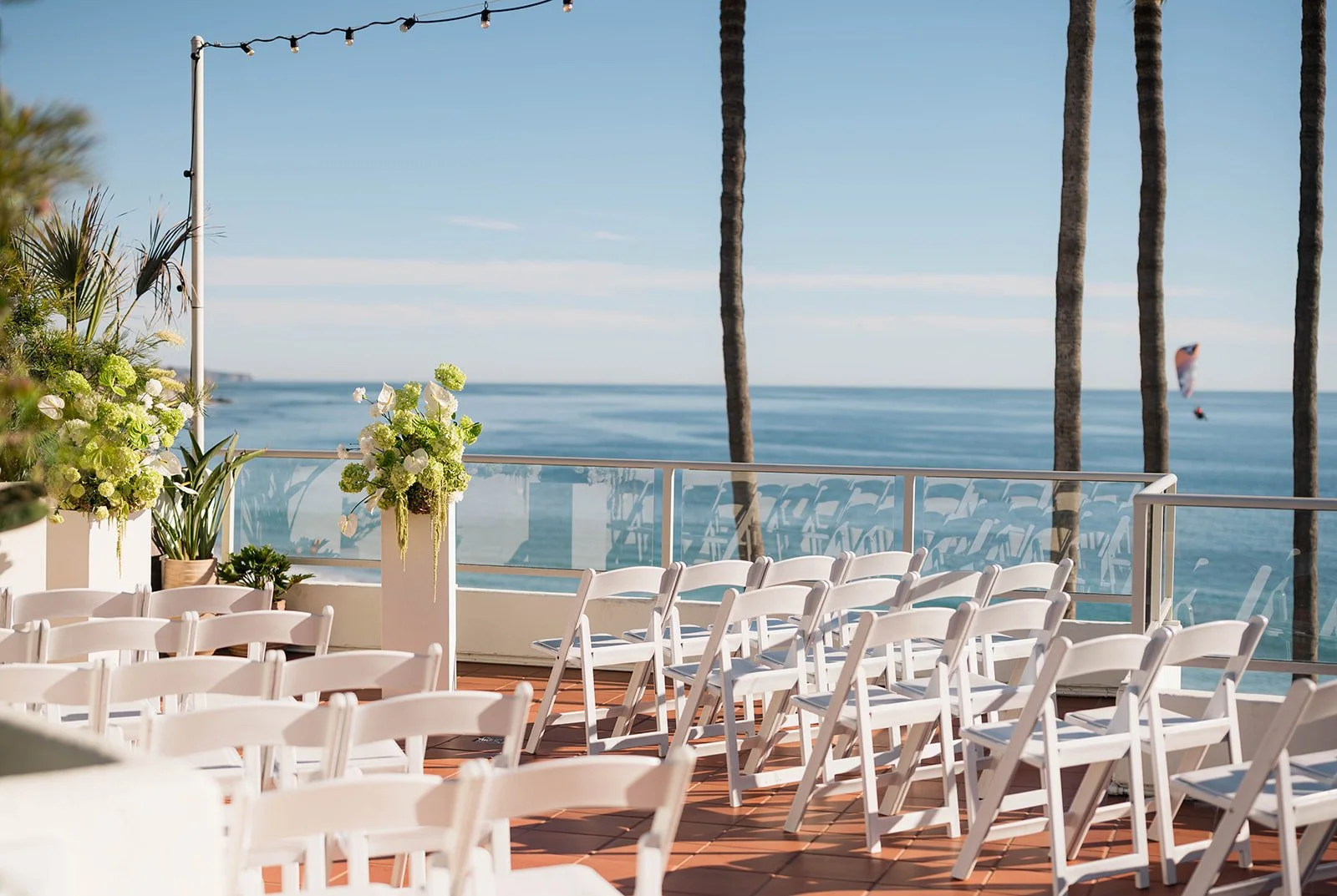 Beachfront ceremony setup with white chairs and green-and-white floral pillars.