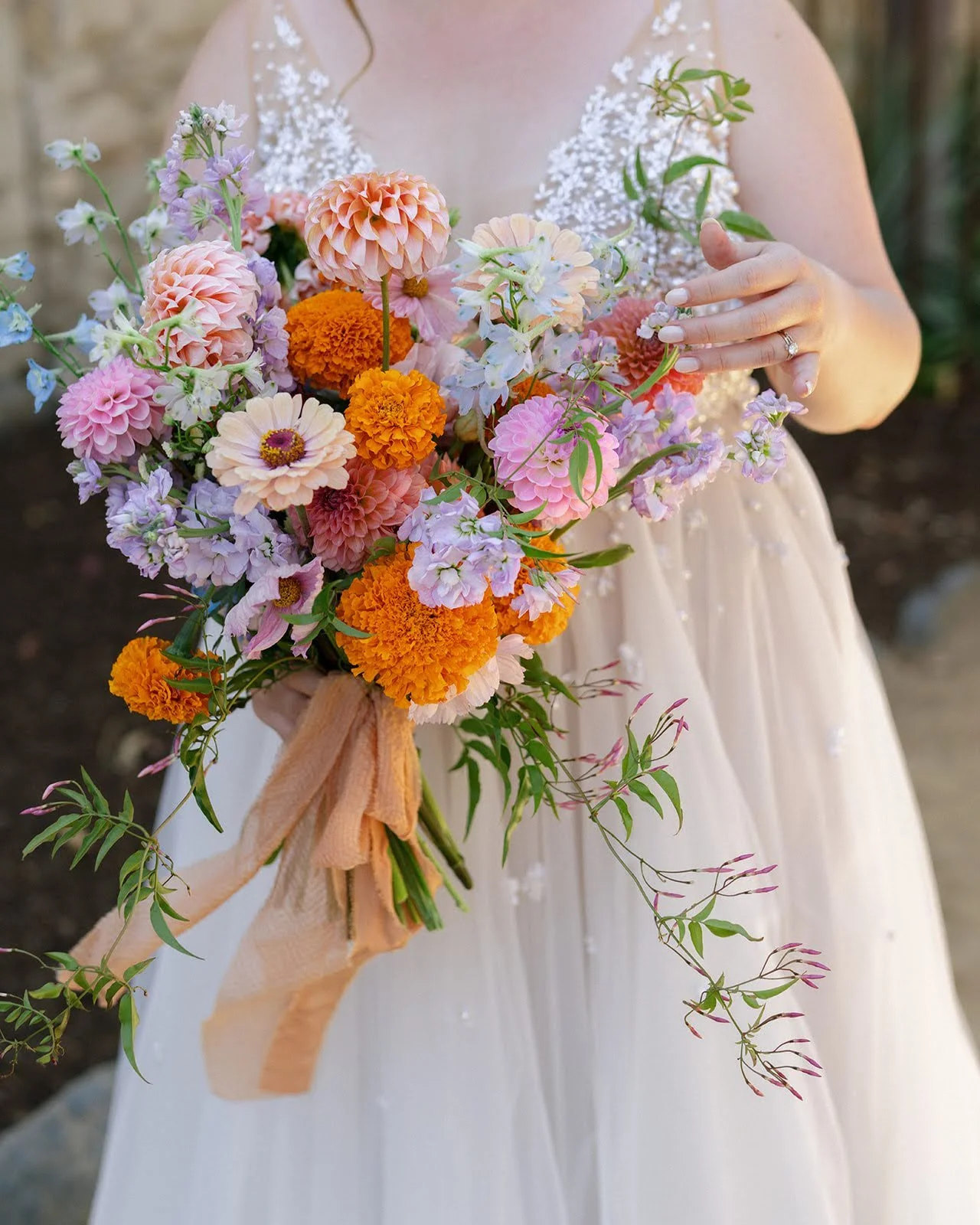 Bride holding a colorful bouquet with dahlias, marigolds, zinnias, and pastel seasonal flowers.