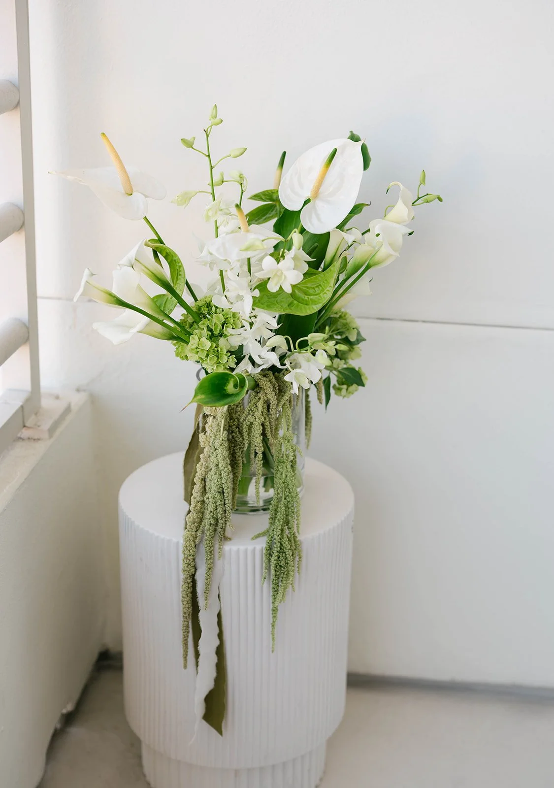 White and green bouquet with anthuriums, calla lilies, orchids, and hanging amaranthus in a glass vase