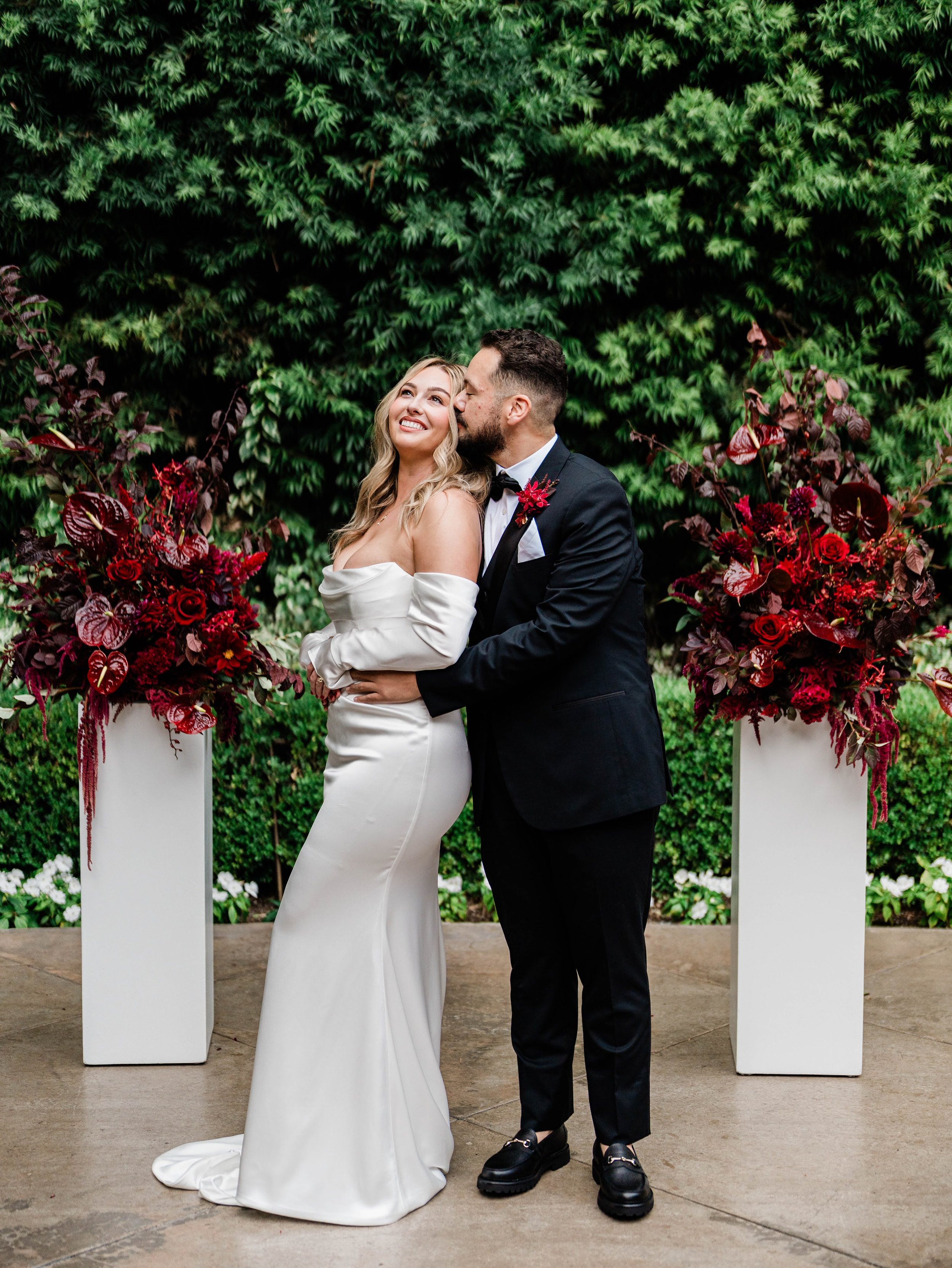 Couple standing between tall burgundy floral pillars during their outdoor ceremony.
