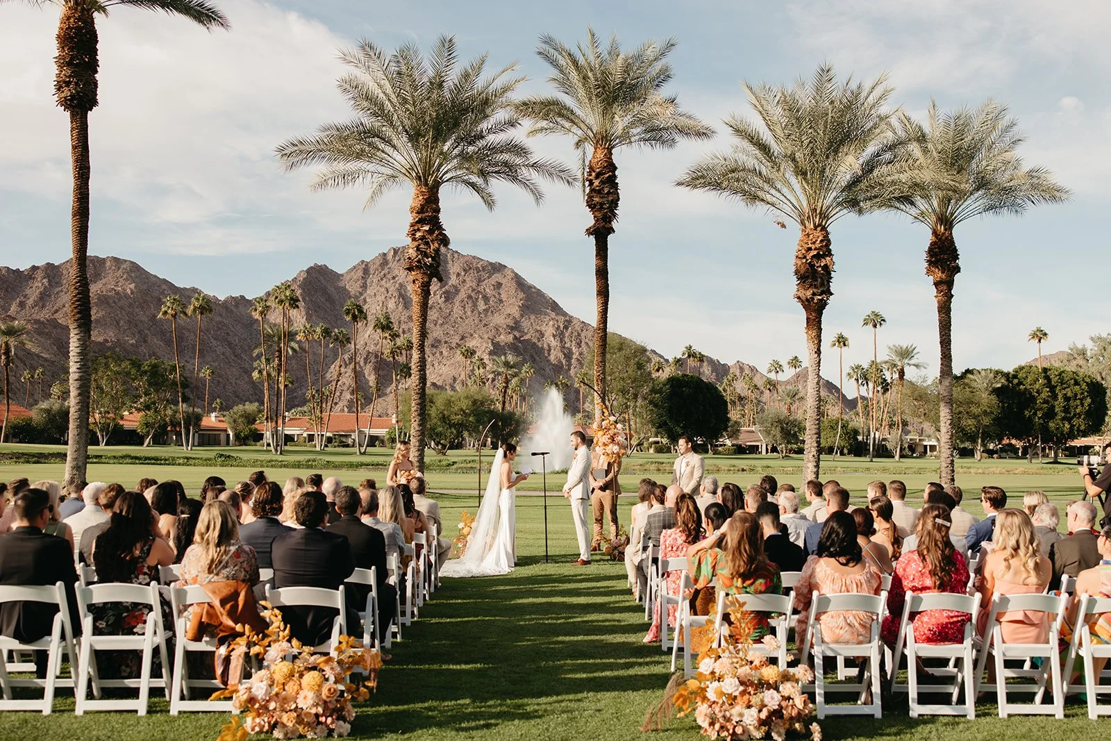 Wide view of a La Quinta Resort wedding ceremony with guests seated along a floral-lined aisle beneath palm trees.