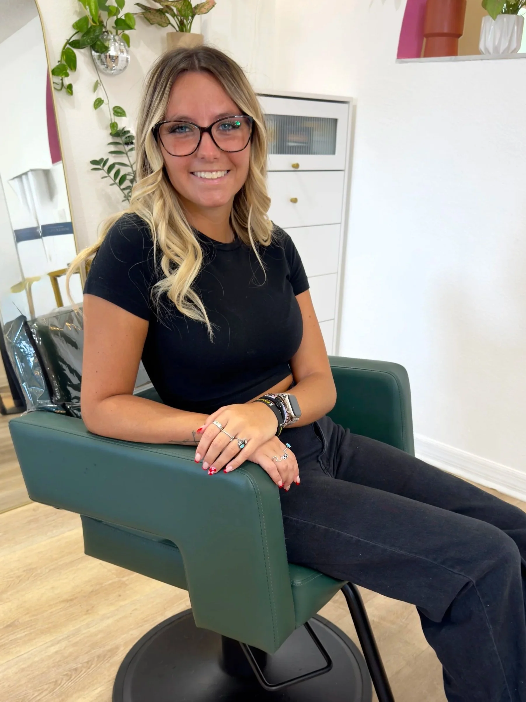 A woman with blonde hair, wearing glasses and a black shirt, sitting in a green chair with her arms crossed, smiling at the camera in a room with plants and white furniture.
