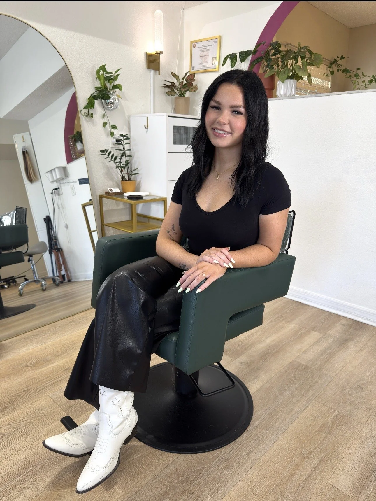 A woman with black hair sitting in a salon chair, wearing a black t-shirt, leather pants, and white cowboy boots. The salon has plants on shelves and a mirror, with a wooden floor.