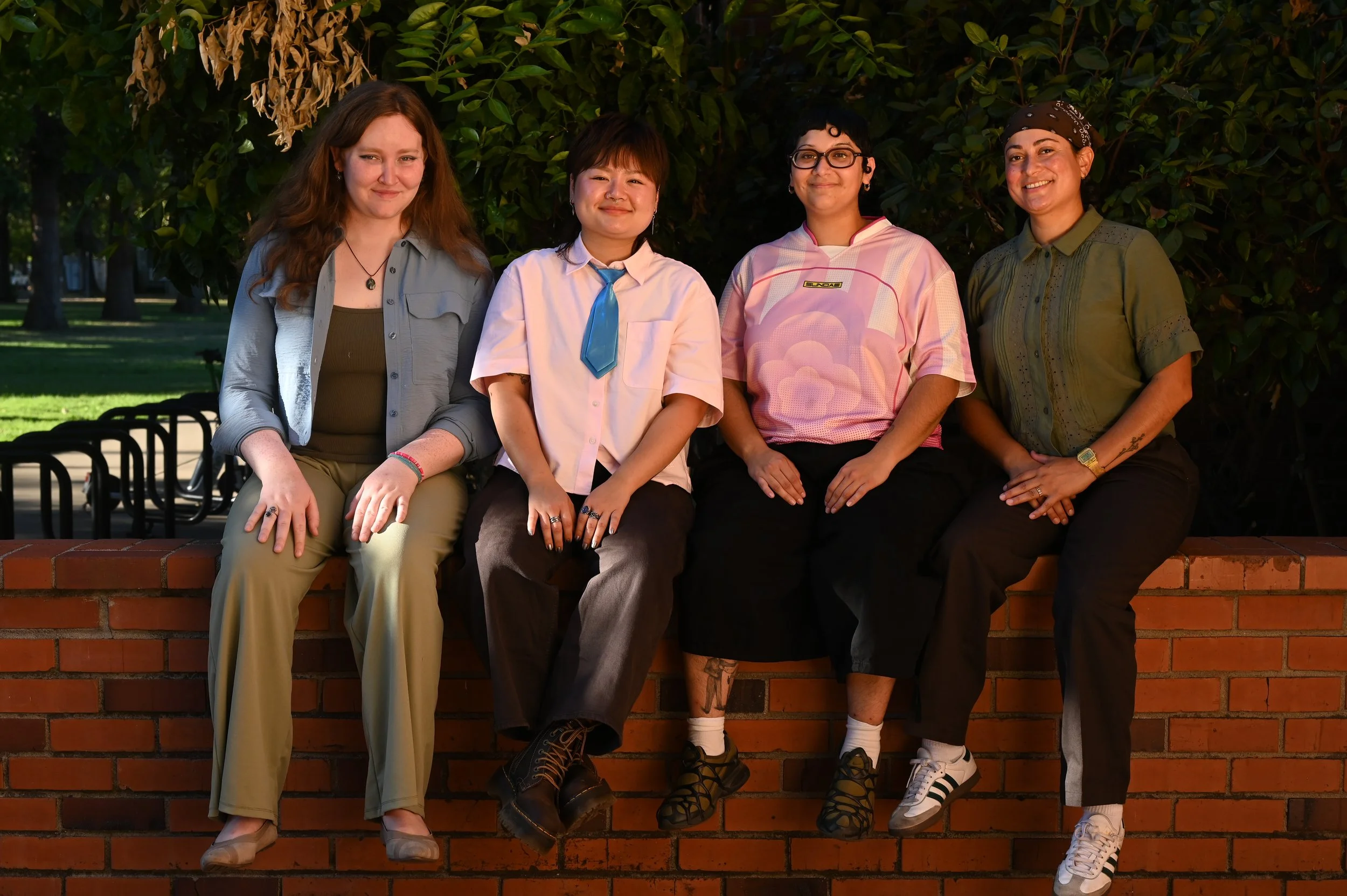 Four people sitting on a brick wall outdoors with green trees in the background, smiling at the camera.