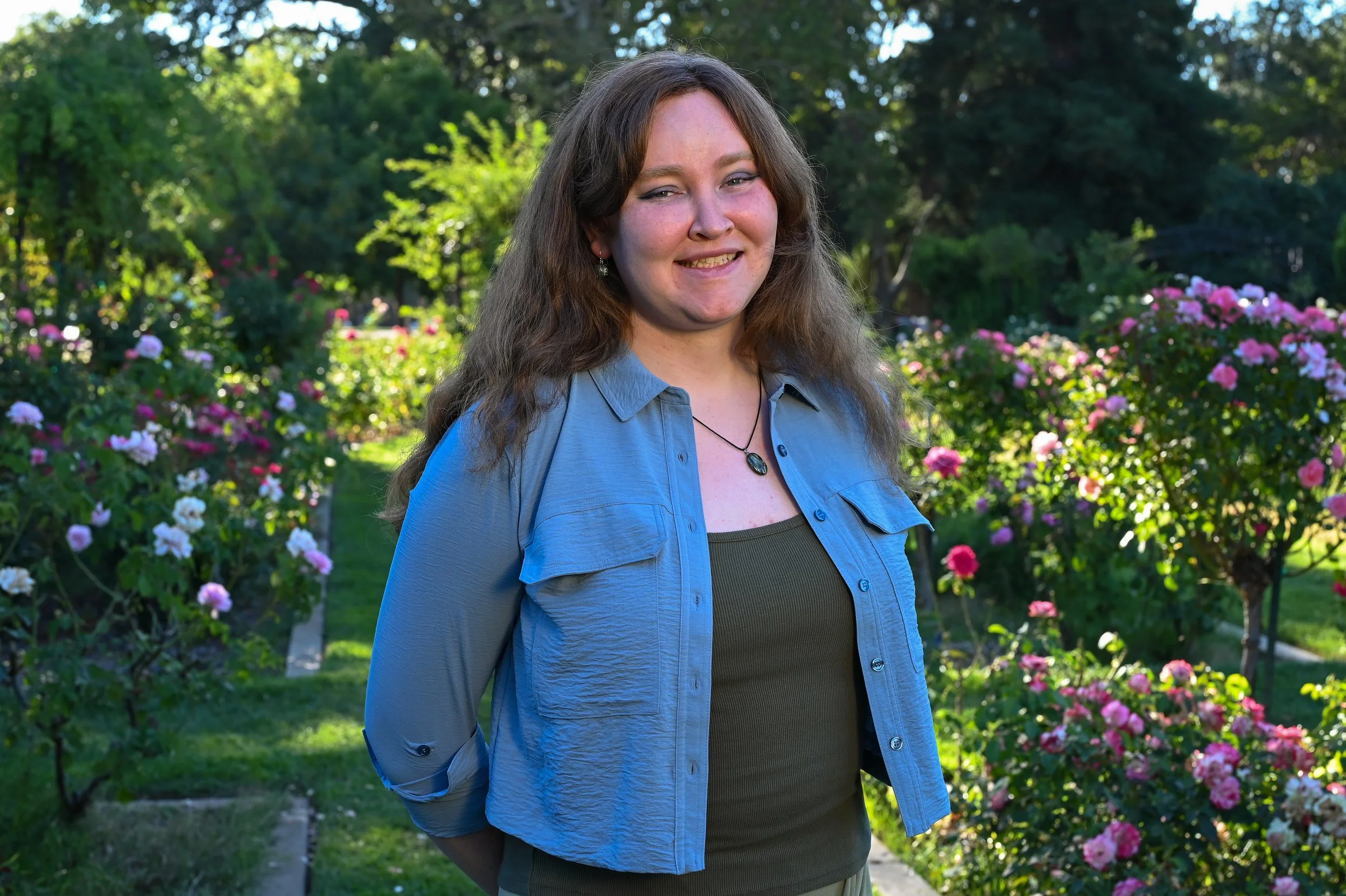 A smiling woman with long brown hair wearing a blue button-up shirt over a green top stands in a garden with blooming pink and white flowers and green trees in the background.