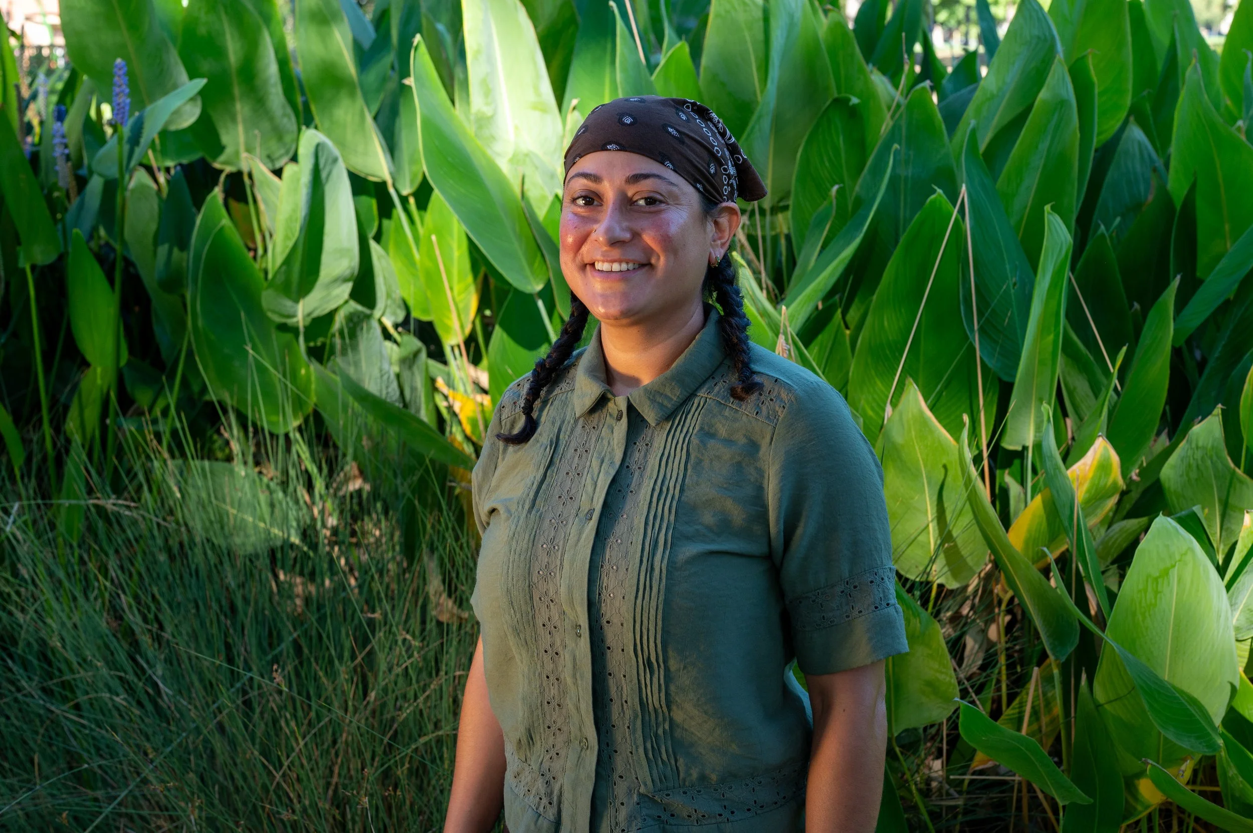 A woman with braided hair, wearing a black bandana and a green shirt, standing outdoors in front of large green leafy plants.