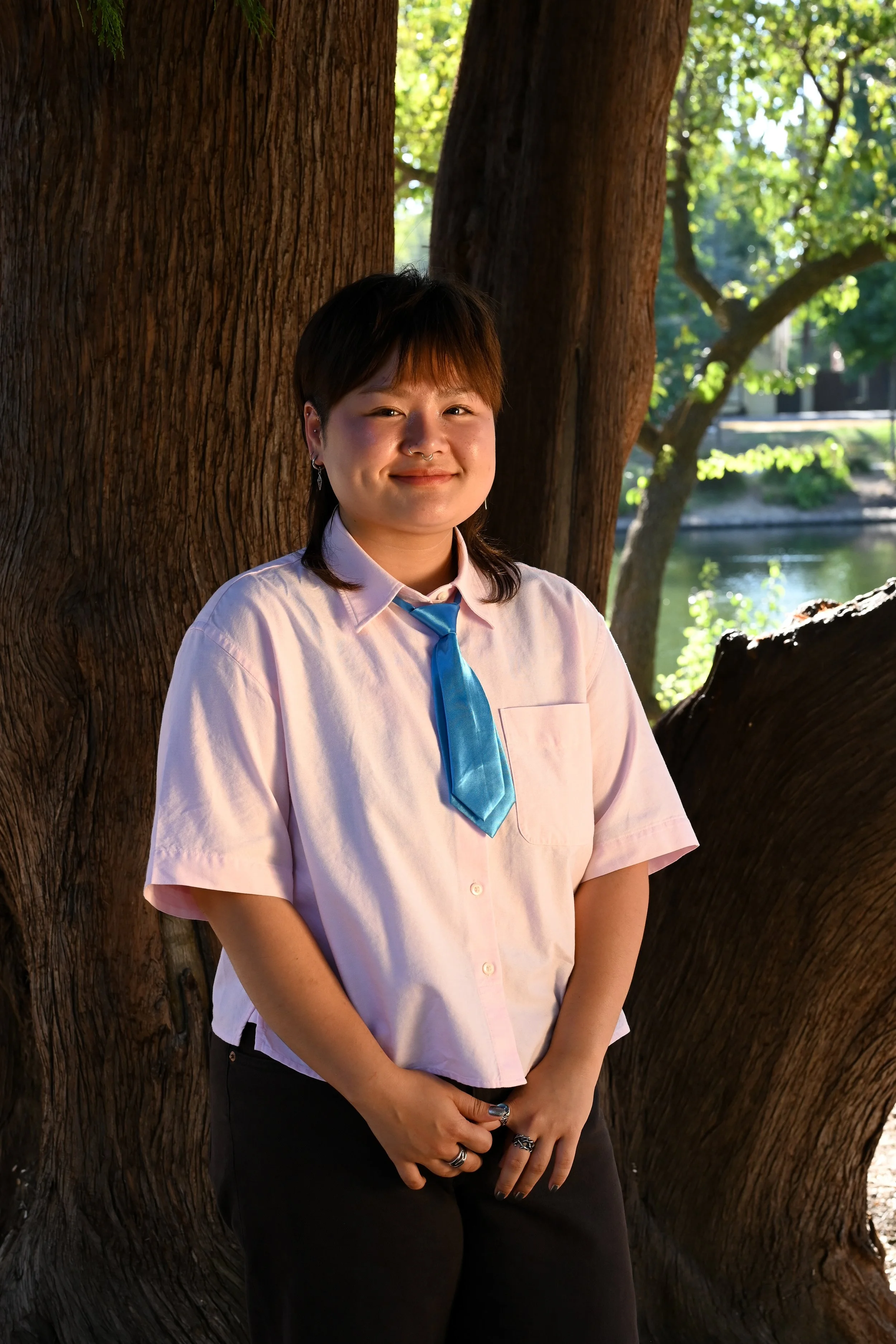 A young person with short dark hair, wearing a light pink button-up shirt and a blue tie, standing outdoors next to a large tree with textured bark. They are smiling gently and standing with hands clasped in front of them, with a background of green trees and a body of water, bathed in warm sunlight.
