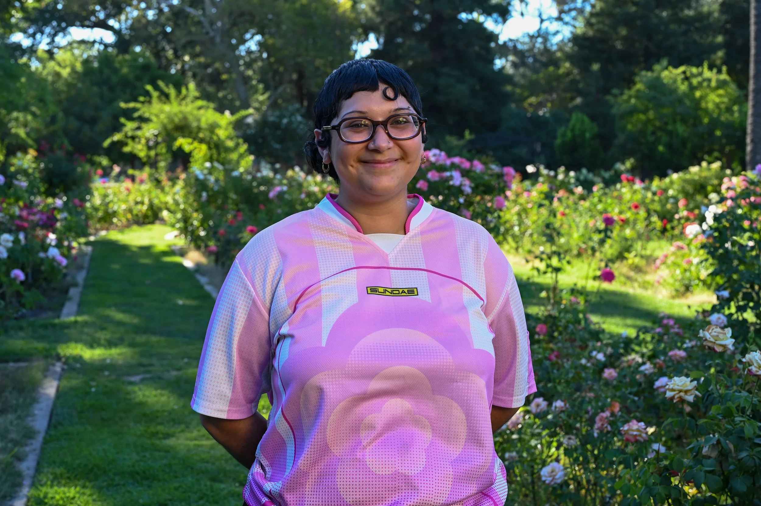 A person with short black hair, glasses, and earrings standing outdoors in a garden with pink flowers and green foliage.