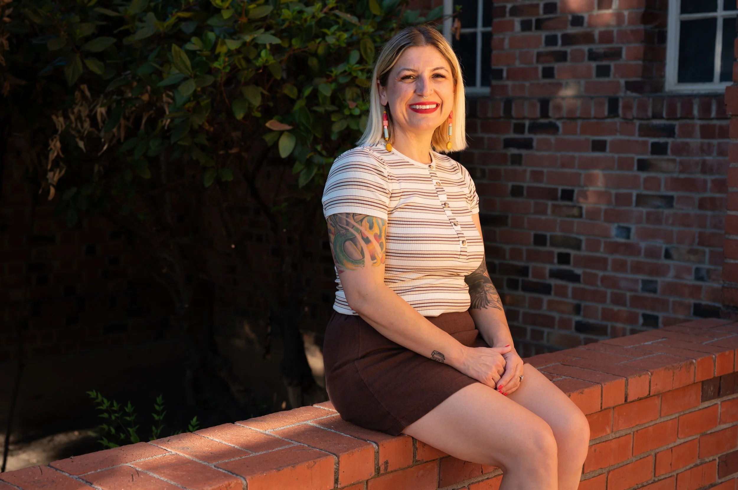 A smiling woman with blonde hair, colorful earrings, and tattoos on her arm, sitting on a brick ledge outdoors during golden hour, with greenery and a brick building in the background.