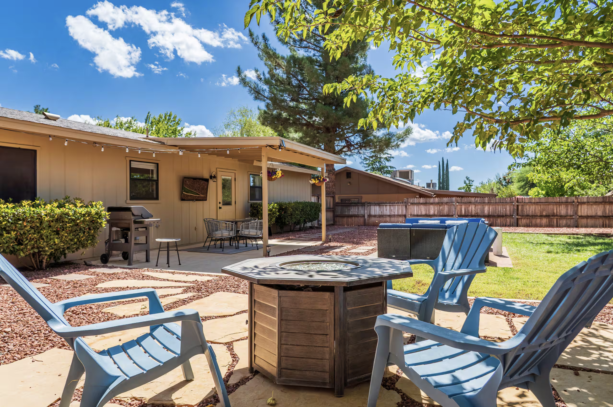 Backyard with outdoor furniture, a patio, a grill, trees, a blue sky with clouds, and a house with string lights.