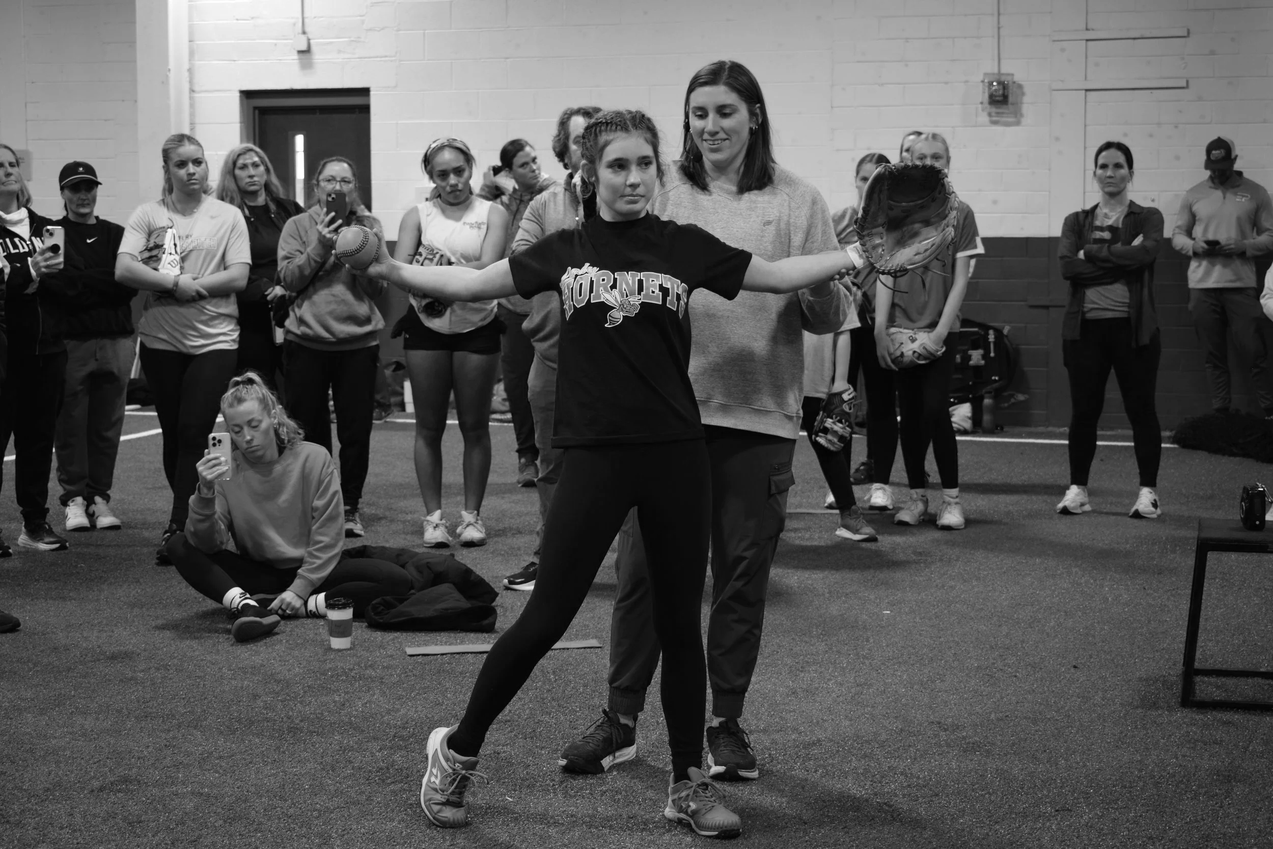A young girl in a sports jersey with "Hornets" printed on it holds a baseball glove and ball, practicing with a coach in an indoor sports facility, with a group of women and girls observing in the background.