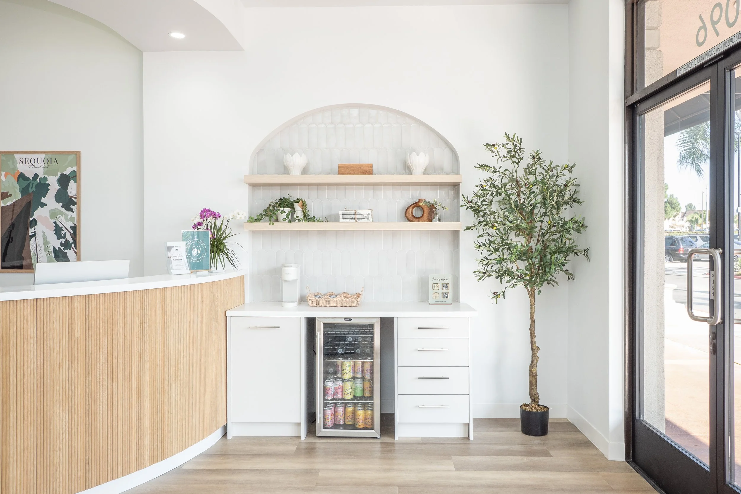 Modern reception area with a white counter, decorative shelves, a small wine cooler with drinks inside, a potted tree, and large glass door with outdoor view.