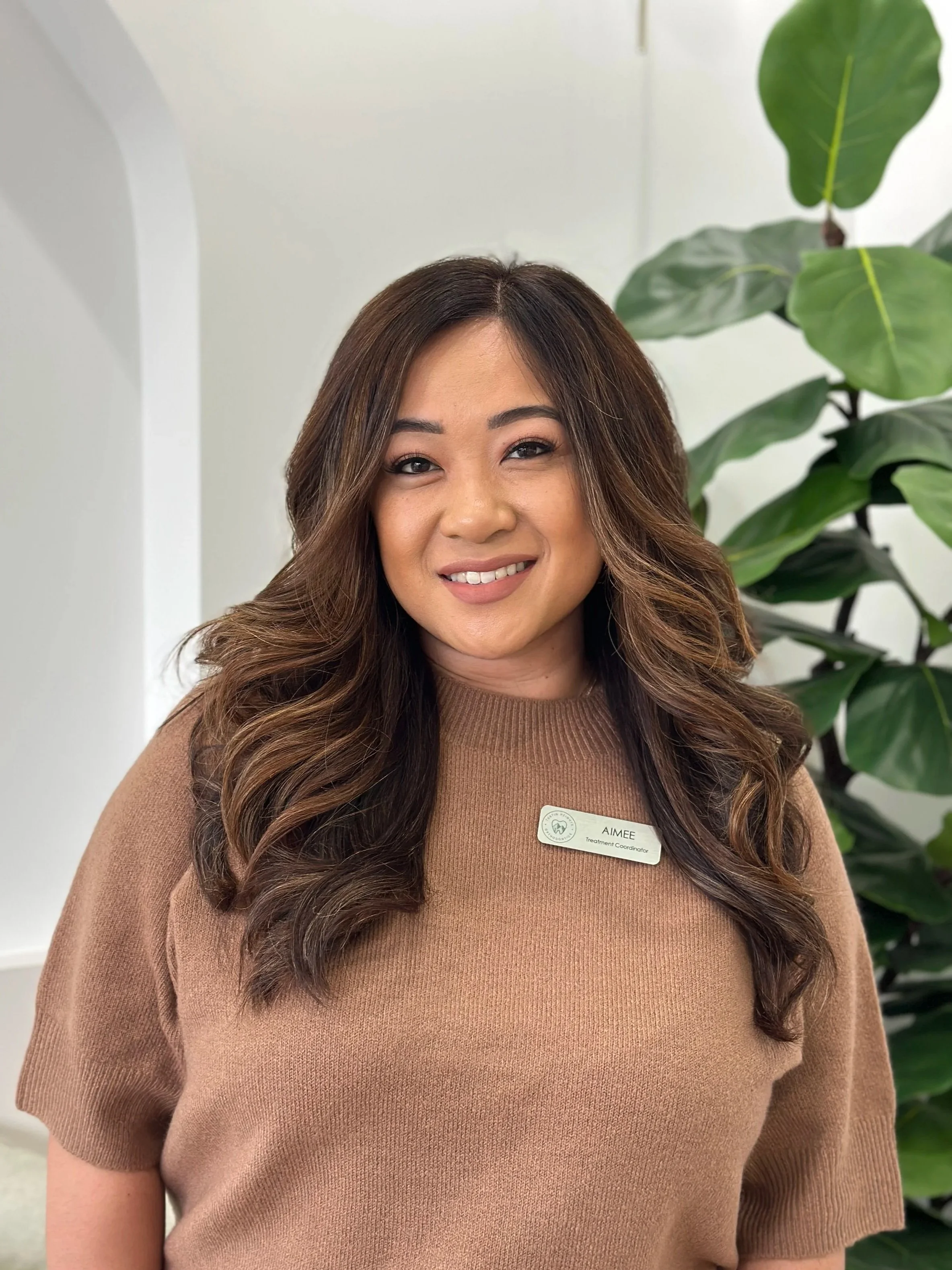 A female with long, brown and wavy hair, smiling and wearing a brown sweater with a silver name tag with the Tustin Heights Orthodontics logo, with a green fiddle leaf fig tree in the background