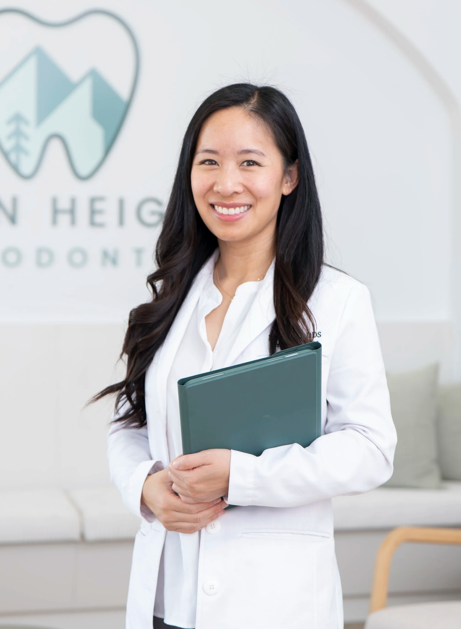 A smiling female healthcare professional in a white coat holding a folder, standing in a modern medical office with a logo that reads 'Linden Heights' in the background.