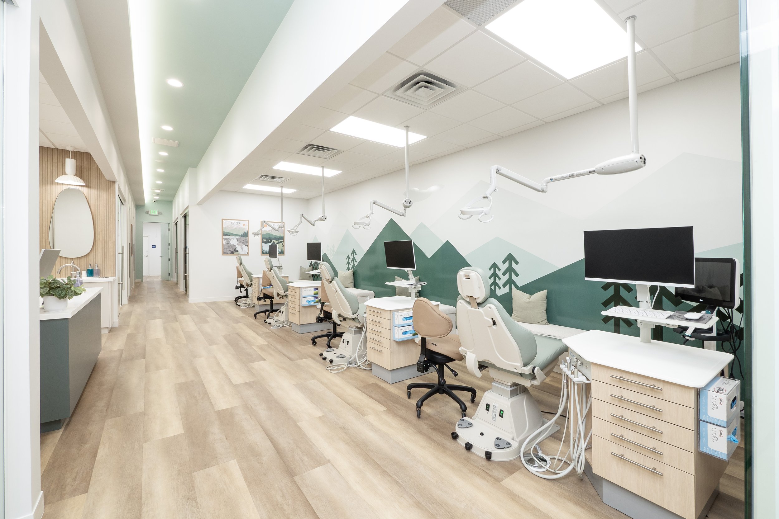 Modern medical treatment room with multiple dental chairs, monitors, and overhead light arms, decorated with a mountain and tree mural mural on the wall, and wood flooring.