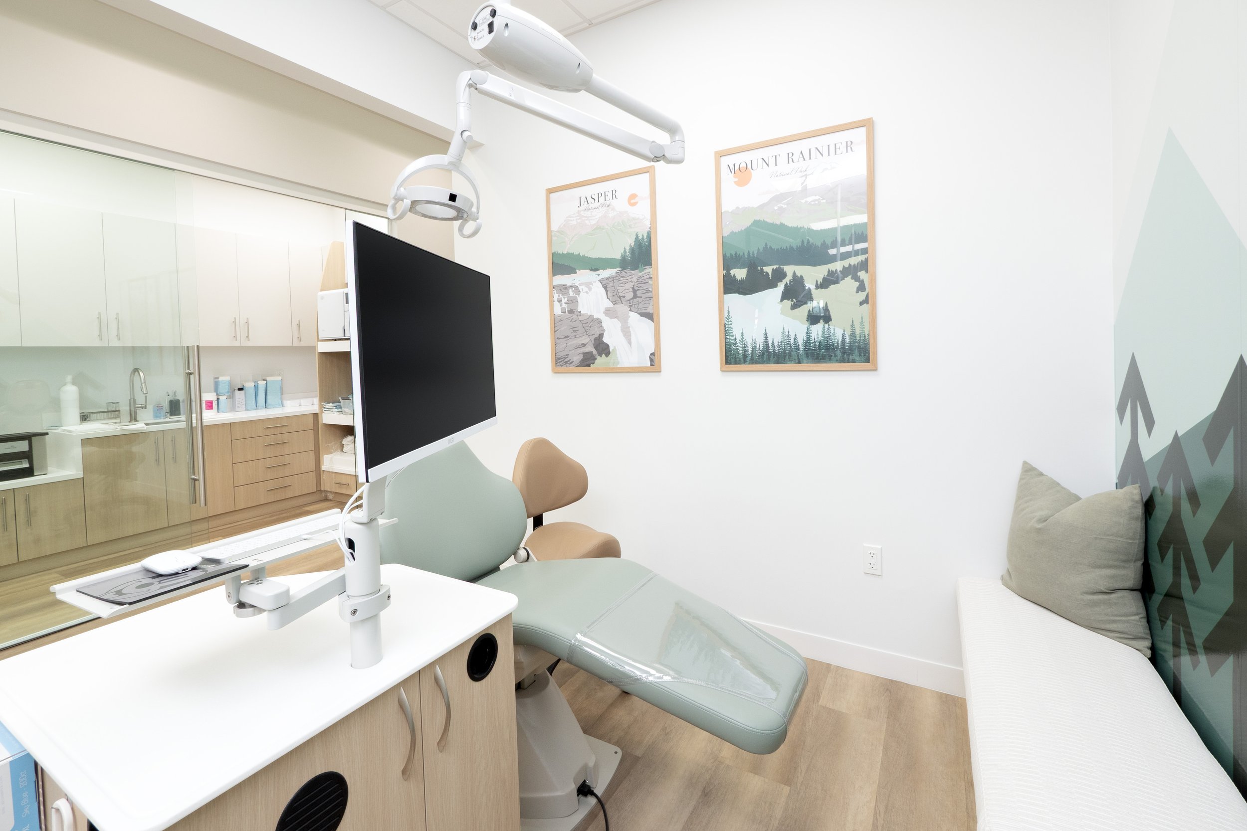 A modern medical examination room with a green examination chair, a wall-mounted monitor, dental or medical equipment, a cabinet, and nature-themed posters on the wall.