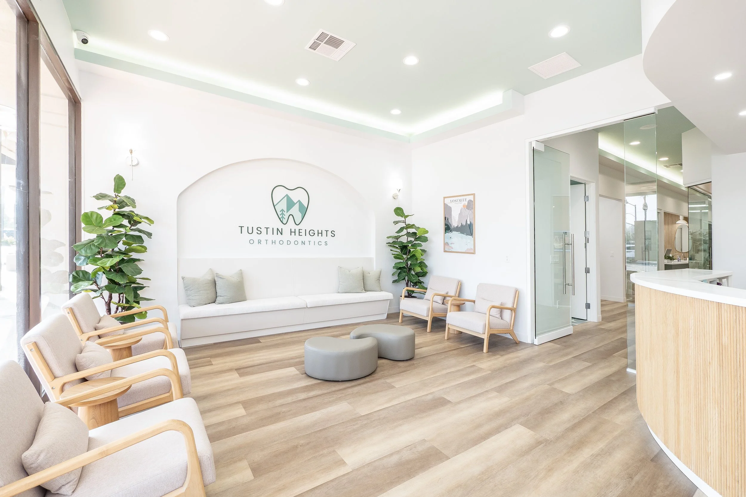 Modern orthodontic office waiting area with white walls, wooden flooring, seating with wooden frames, plants, and a sign that reads "Tustin Heights Orthodontics."