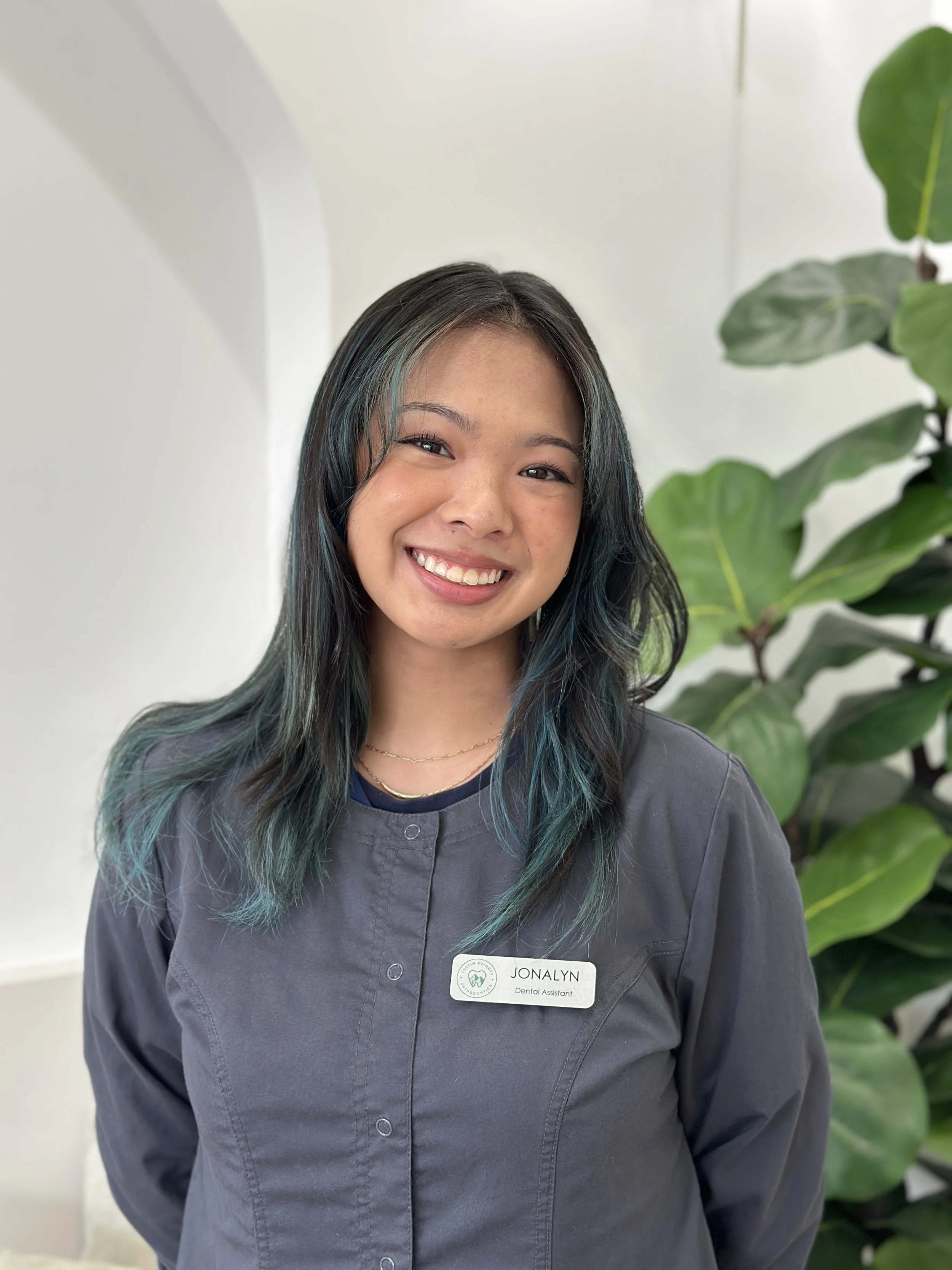 A female with long, black and blue straight hair, smiling and wearing a pewter grey jacket with a silver name tag with the Tustin Heights Orthodontics logo, with a green fiddle leaf fig tree in the background
