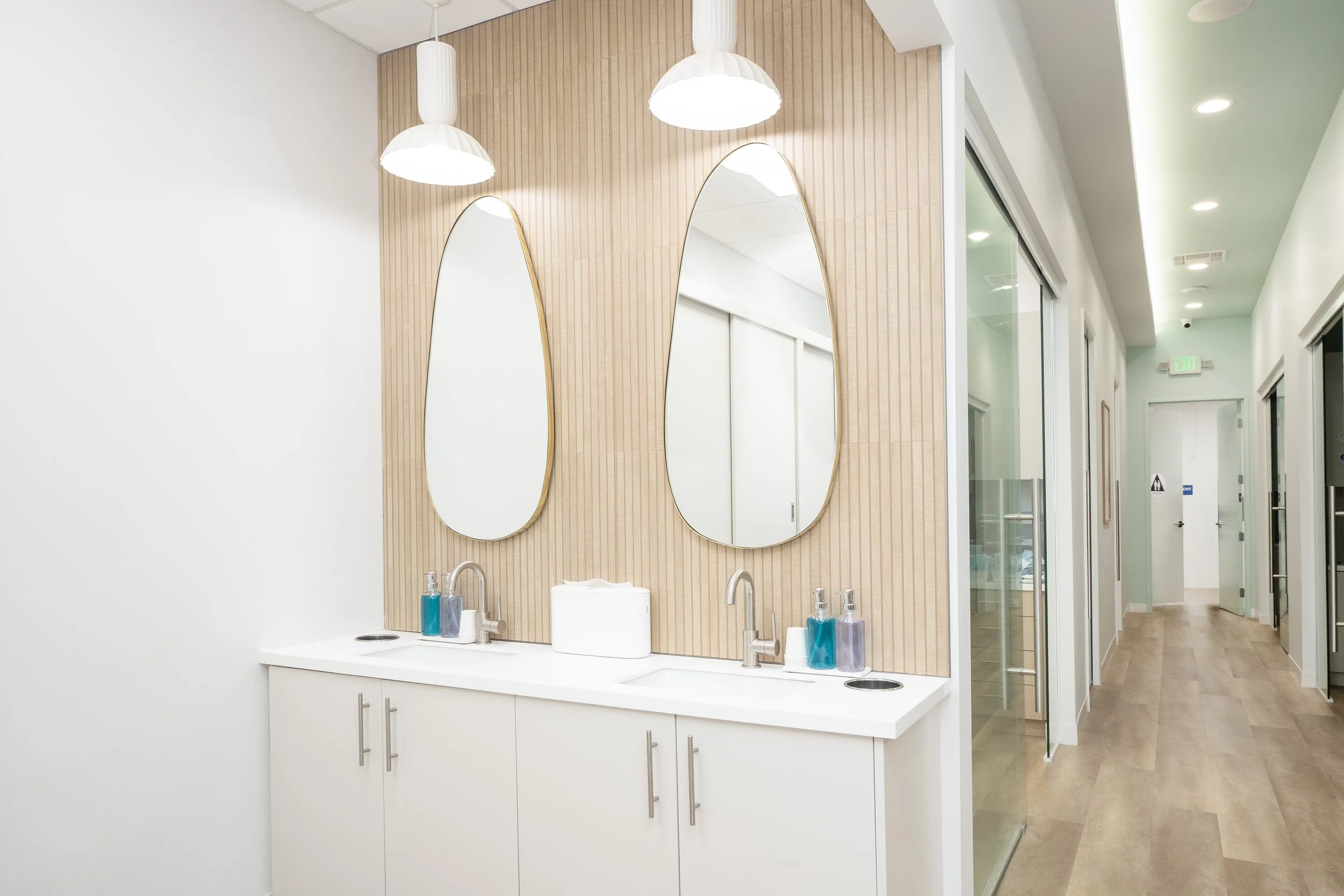 Modern bathroom with two oval mirrors, double sinks, white cabinetry, beige wall paneling, pendant lights, and a hallway with wooden flooring and glass doors.