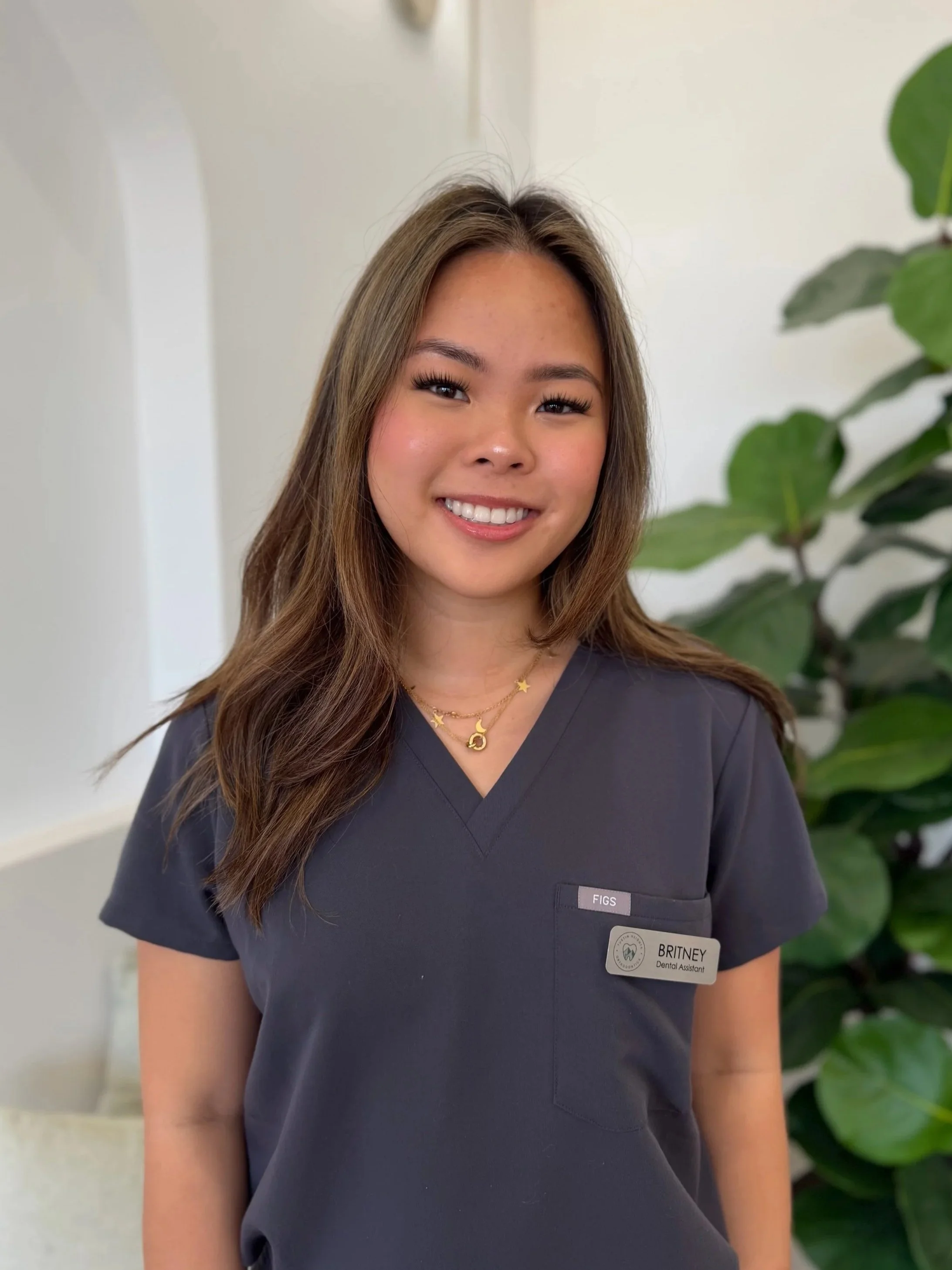 A young woman with long brown hair, smiling, wearing a dark gray medical scrubs with name tag, standing indoors near a green plant.