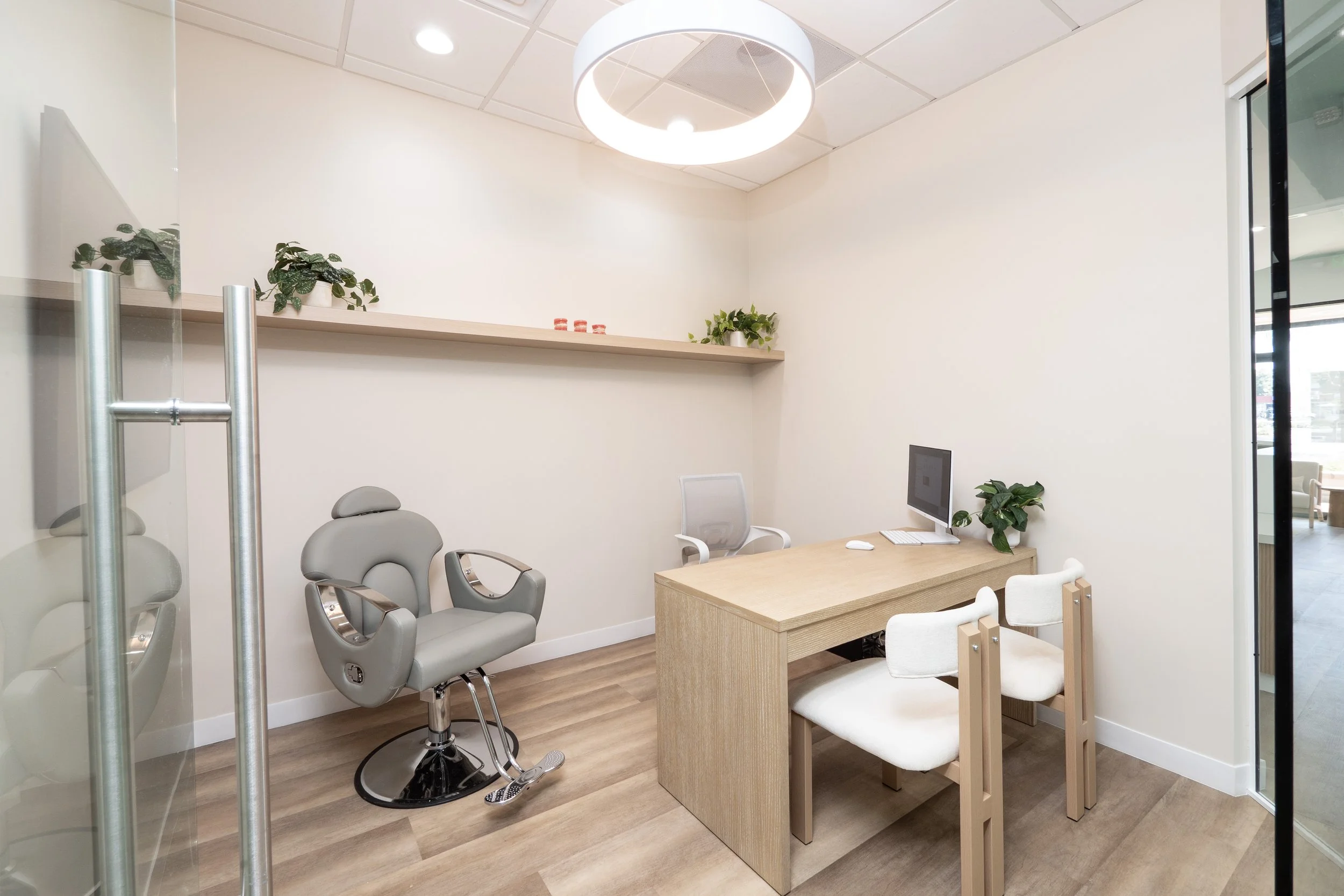 Empty medical consultation room with a grey examination chair, a wooden desk with a computer, white chairs, and decorative plants.