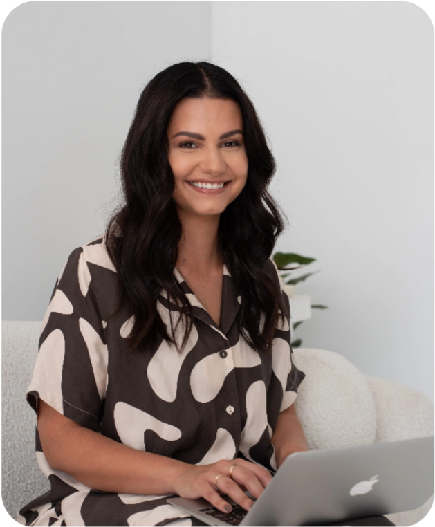 A smiling woman with long dark hair sitting on a light-colored couch, using a silver laptop in a bright, minimalistic room with a white wall and a plant in the background.