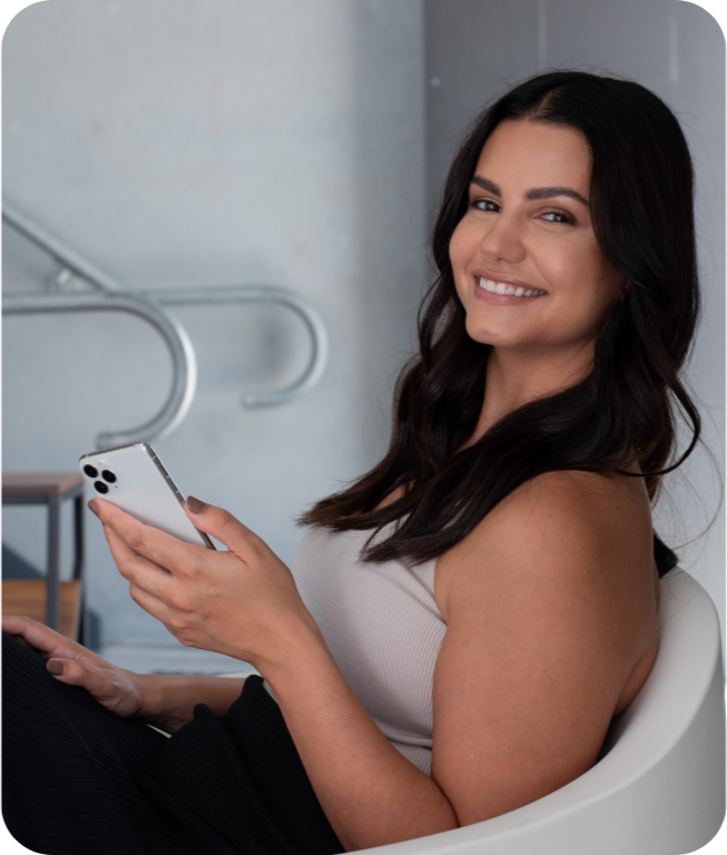 A woman with long dark hair smiling while holding a smartphone in her hand in an indoor setting.