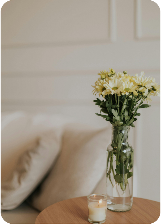 A glass vase with white and yellow flowers on a wooden table next to a small candle, with a bed and pillows in the background.