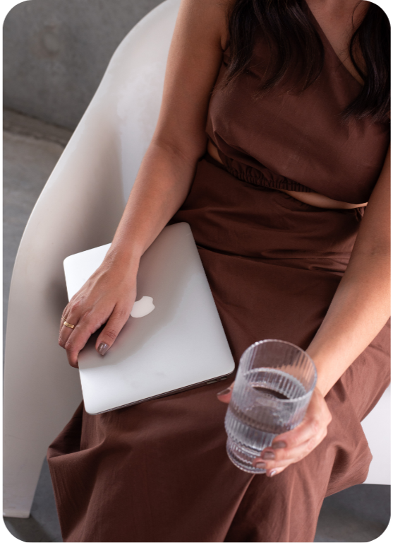 A woman sitting on a white chair holding a glass of water in one hand and a closed silver MacBook in the other, wearing a brown dress.