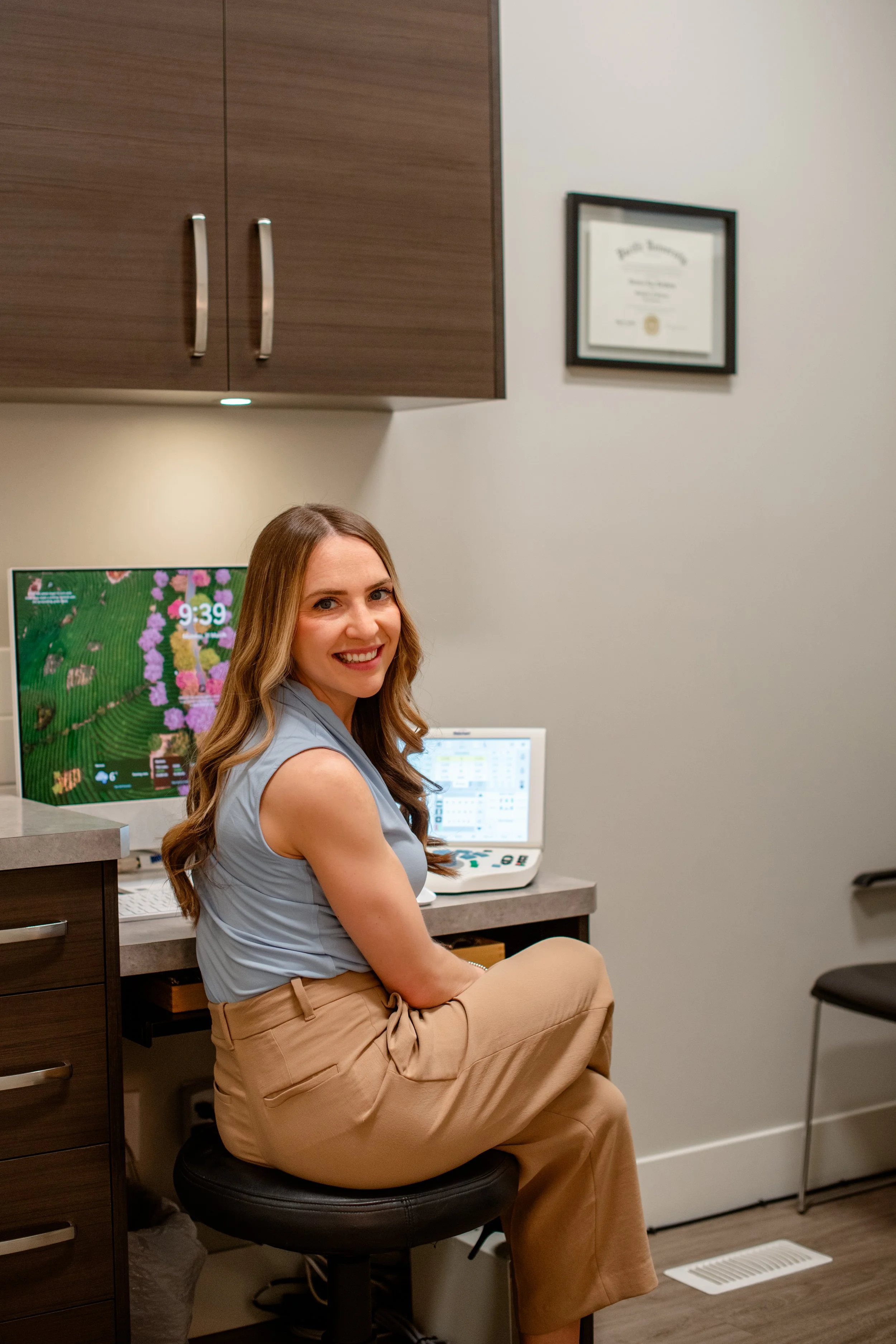 A smiling woman with long brown hair sitting on an office stool in a medical office, with a computer monitor displaying a colorful image and a framed diploma on the wall.