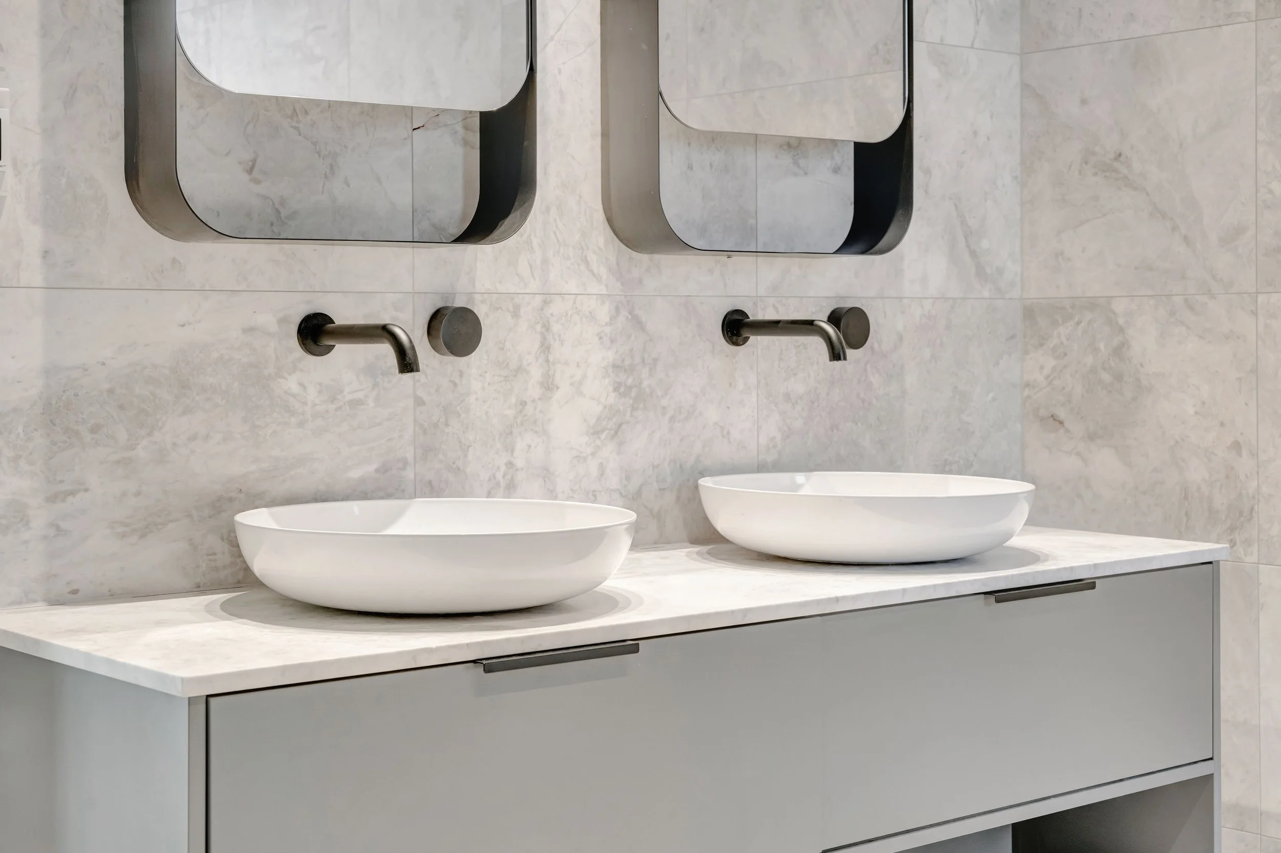 Two white oval bathroom sinks on a marble countertop with two black wall-mounted faucets and two rectangular mirrors above, against a marble wall.