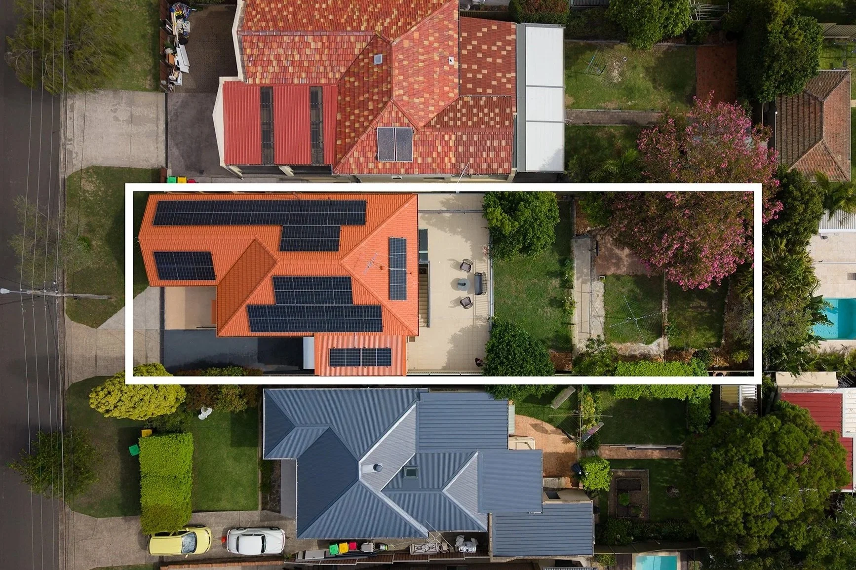An aerial view of residential houses with red and blue roofs, including solar panels, surrounded by green trees and yards, with parked cars on the street and in driveways.