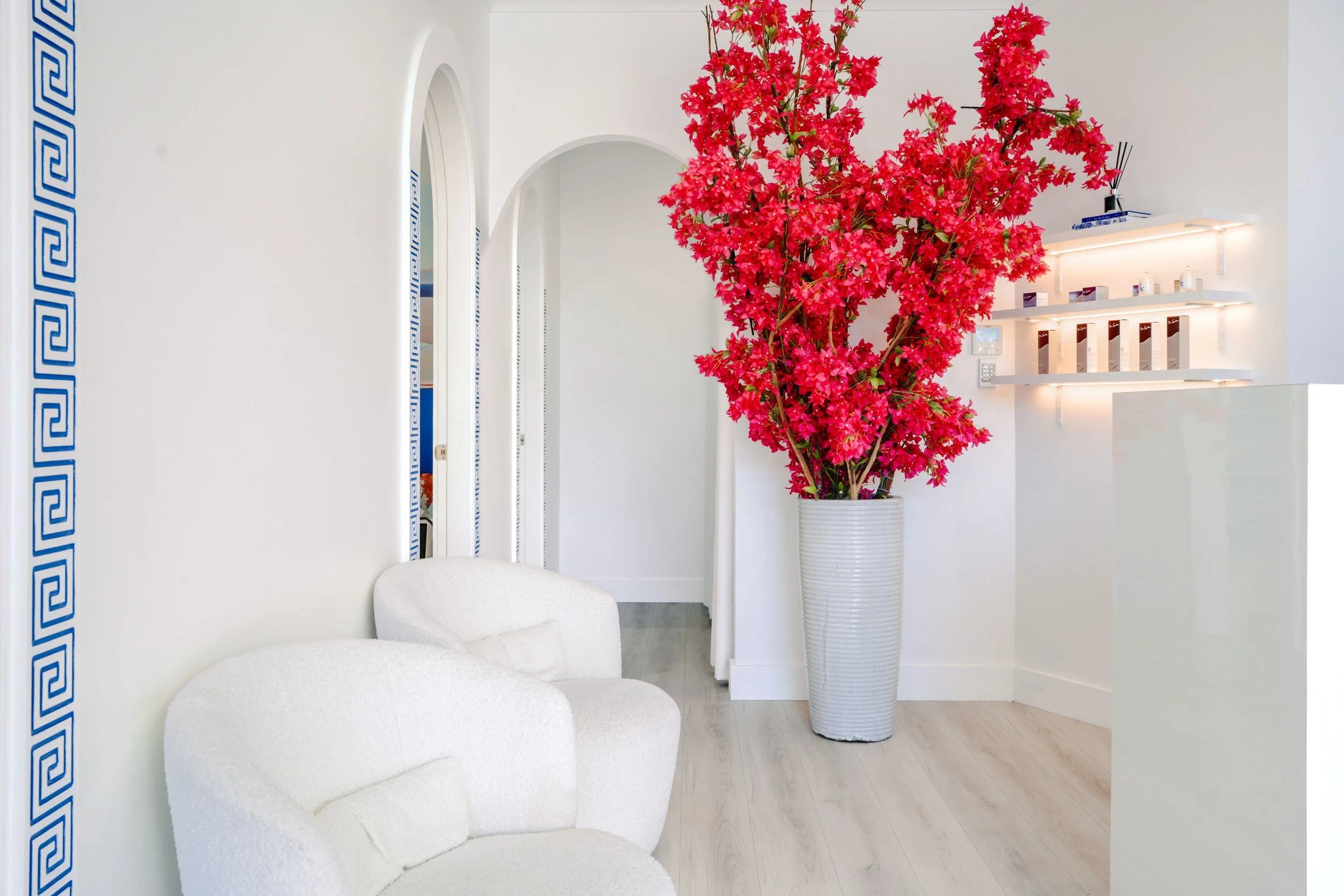 Interior of a modern minimalistic lobby with white walls, white cushioned chairs, and a large pink flowering plant in a tall white vase. There are built-in shelves with small items on the wall and a reception desk to the right.