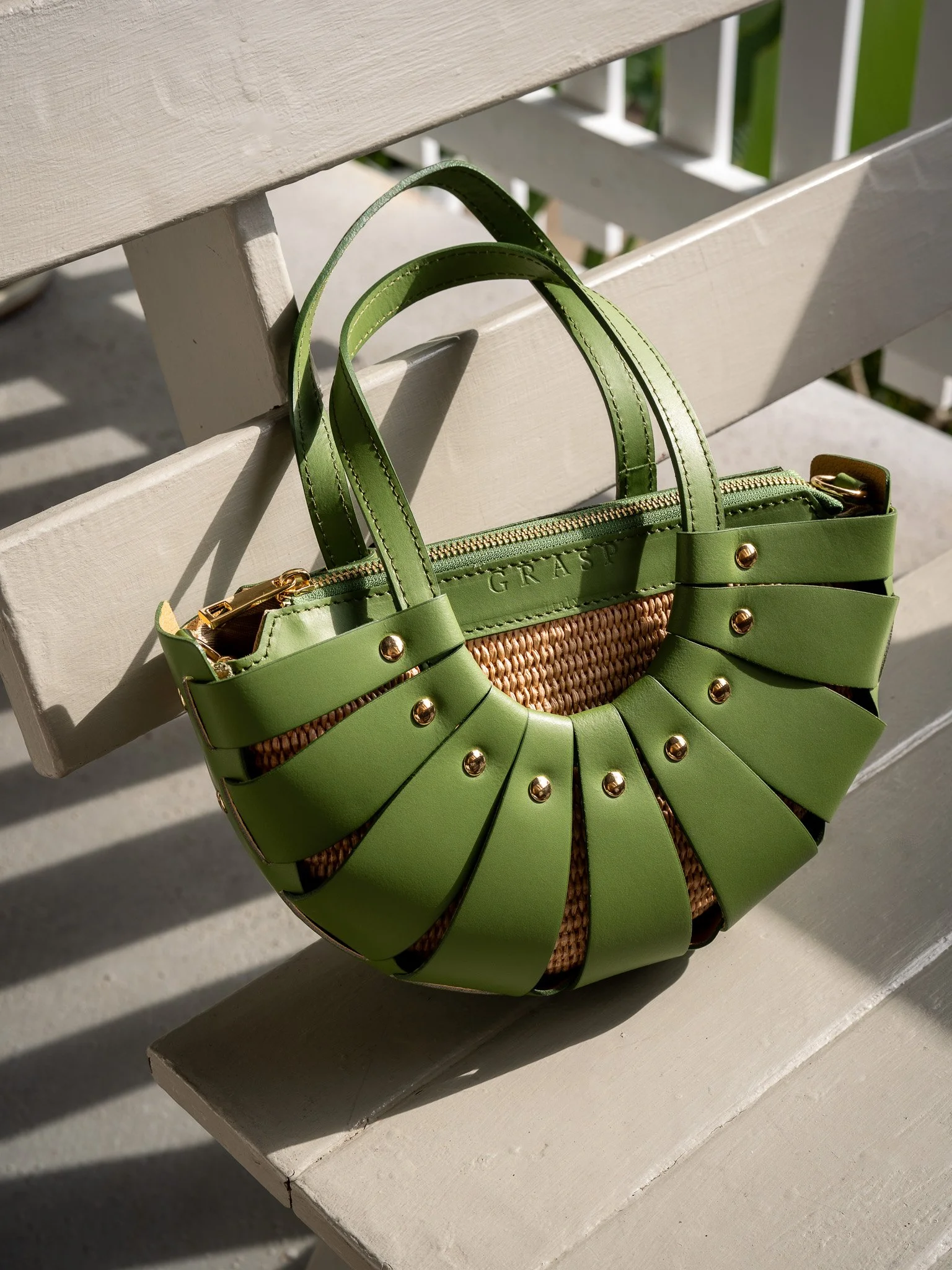A green handbag with gold accents and a woven center, resting on a white wooden bench with sunlight casting shadows.