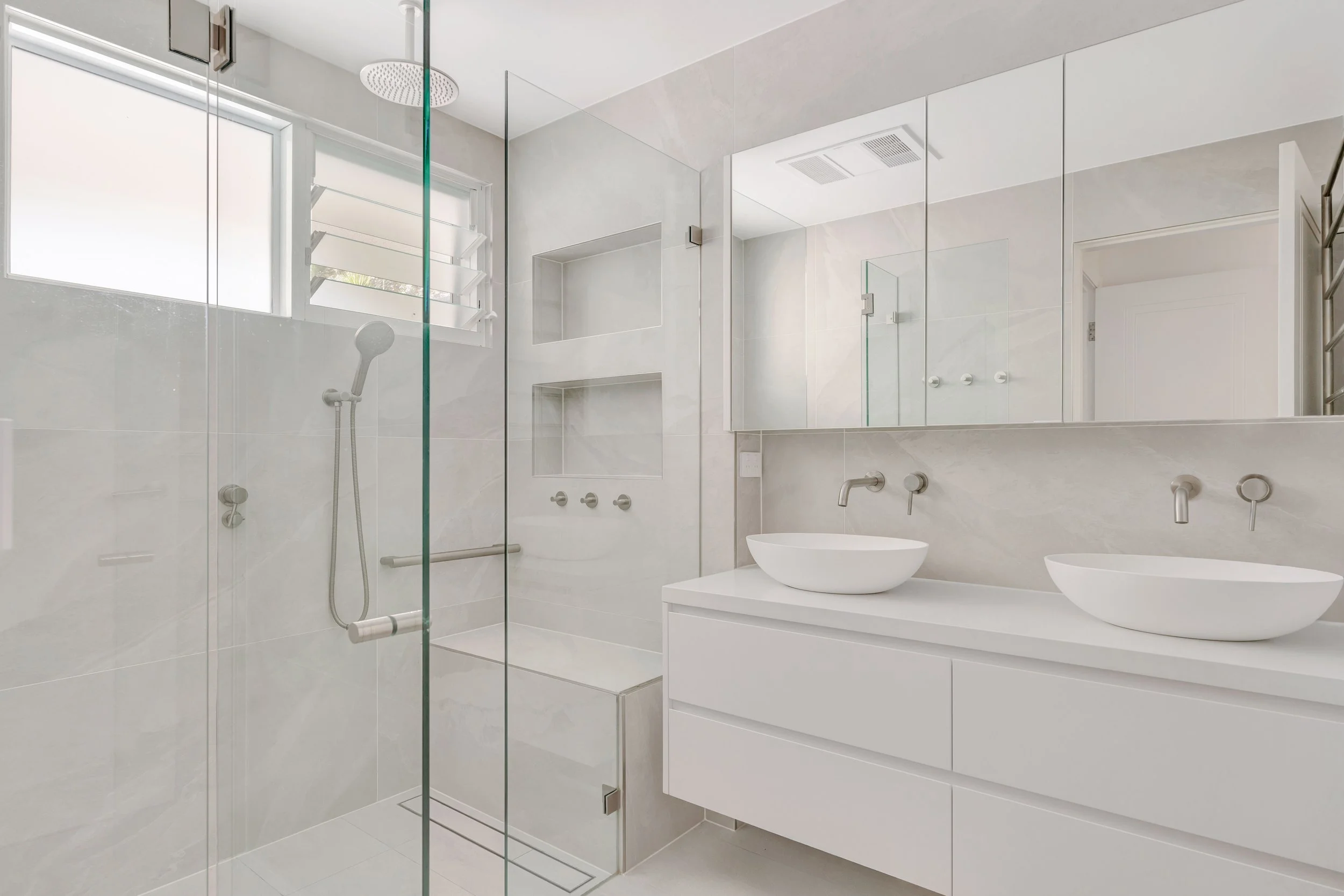 Modern bathroom with walk-in shower, double vanity with vessel sinks, large mirror, window with louvers, and minimalist design.