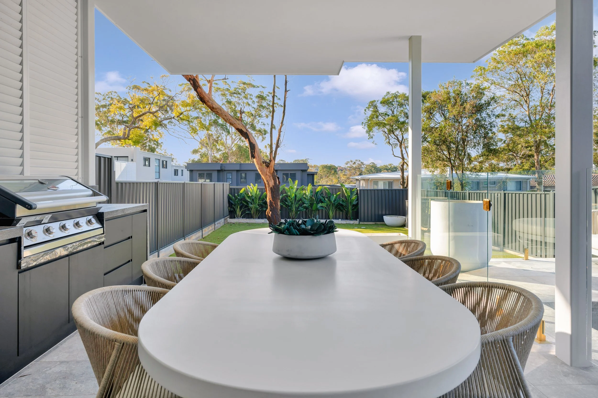 Outdoor patio with a long white dining table, woven chairs, a decorative plant in a bowl, a built-in grill, and a view of a backyard with trees and a fence.