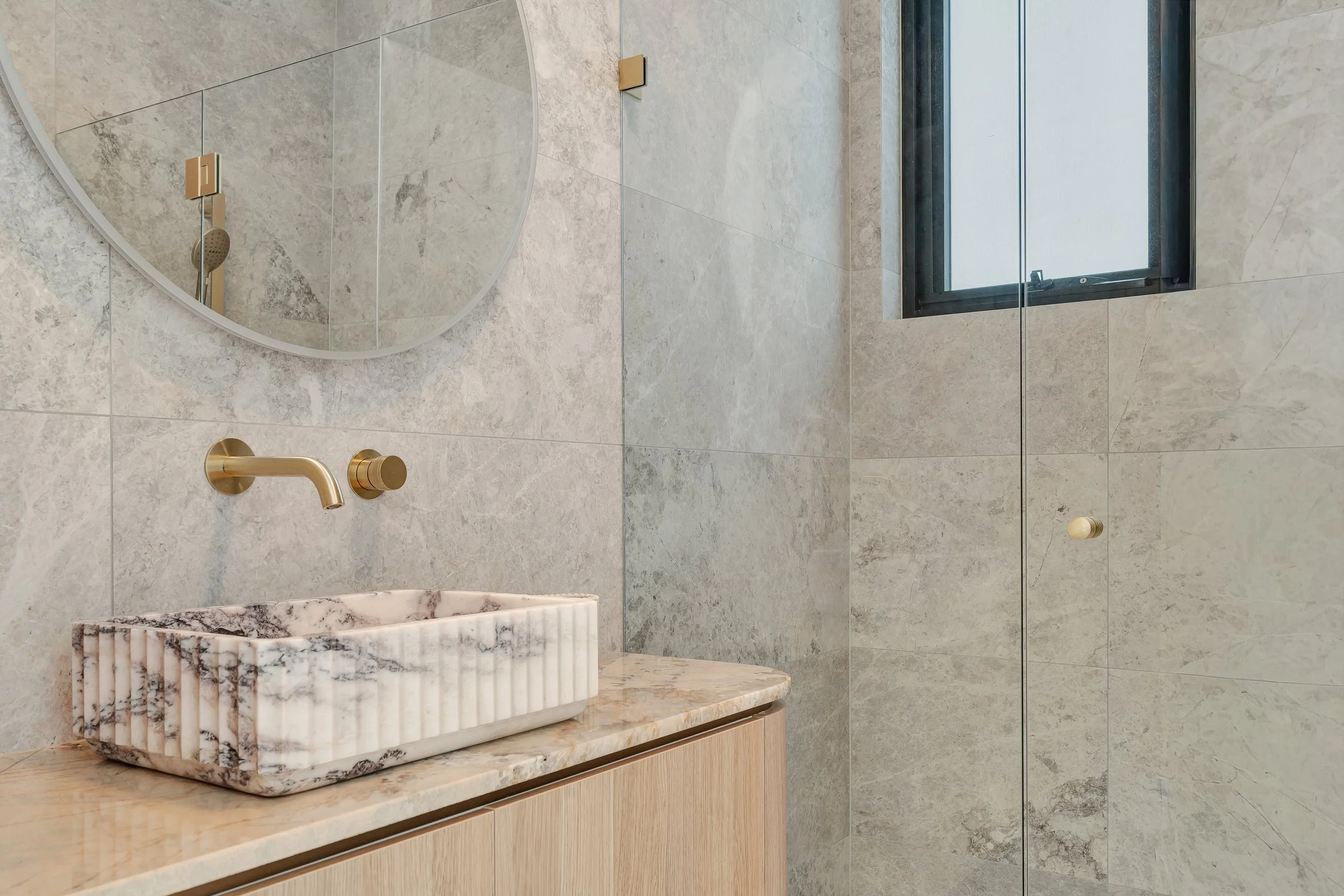 Bathroom with beige marble sink, round mirror, brass faucet, textured tile walls, and a small window