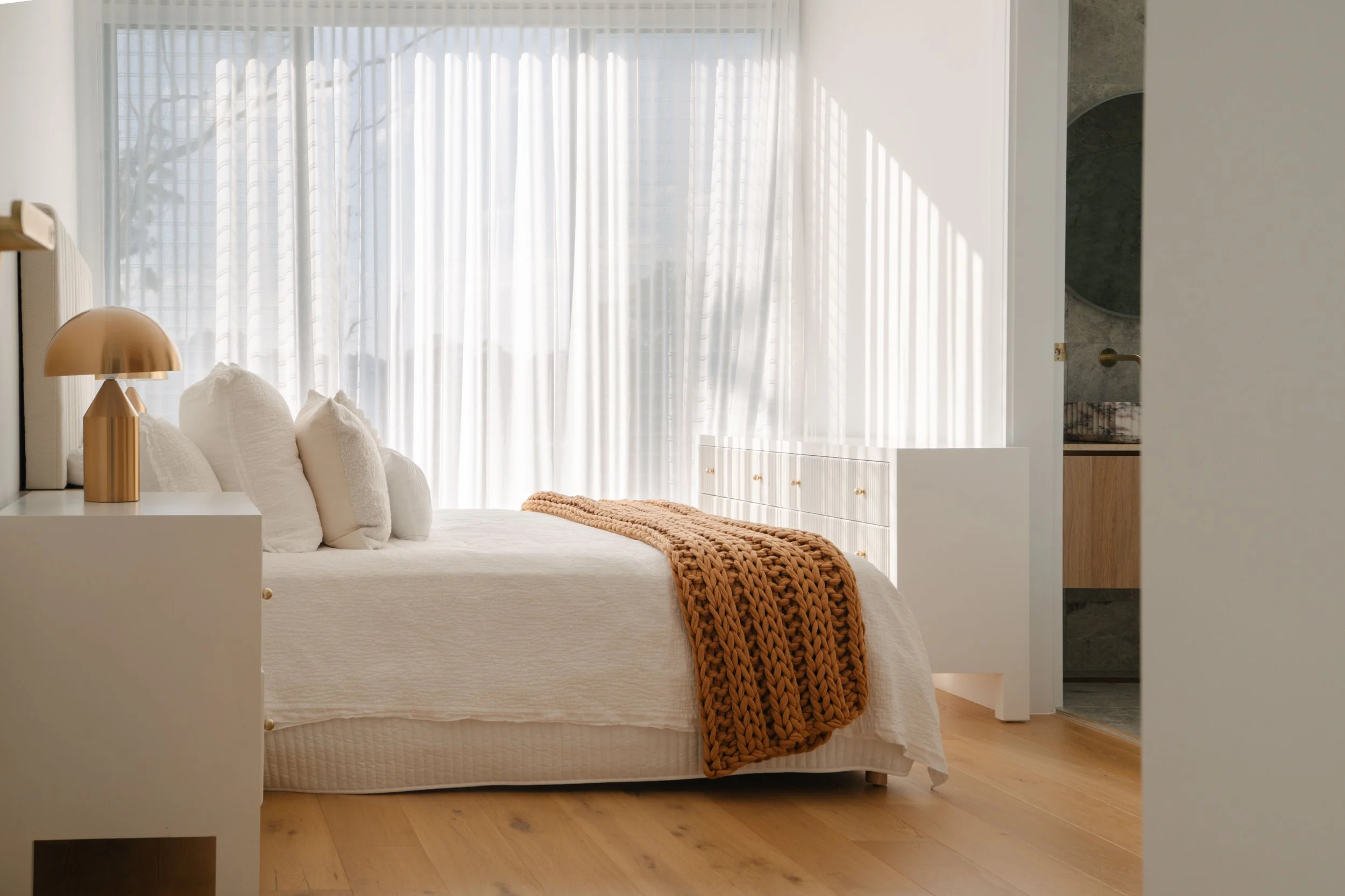 A modern bedroom with a white bed, multiple pillows, a brown chunky knit blanket, a white nightstand with a gold lamp, large windows with sheer white curtains, and a glimpse of a bathroom with a marble sink.