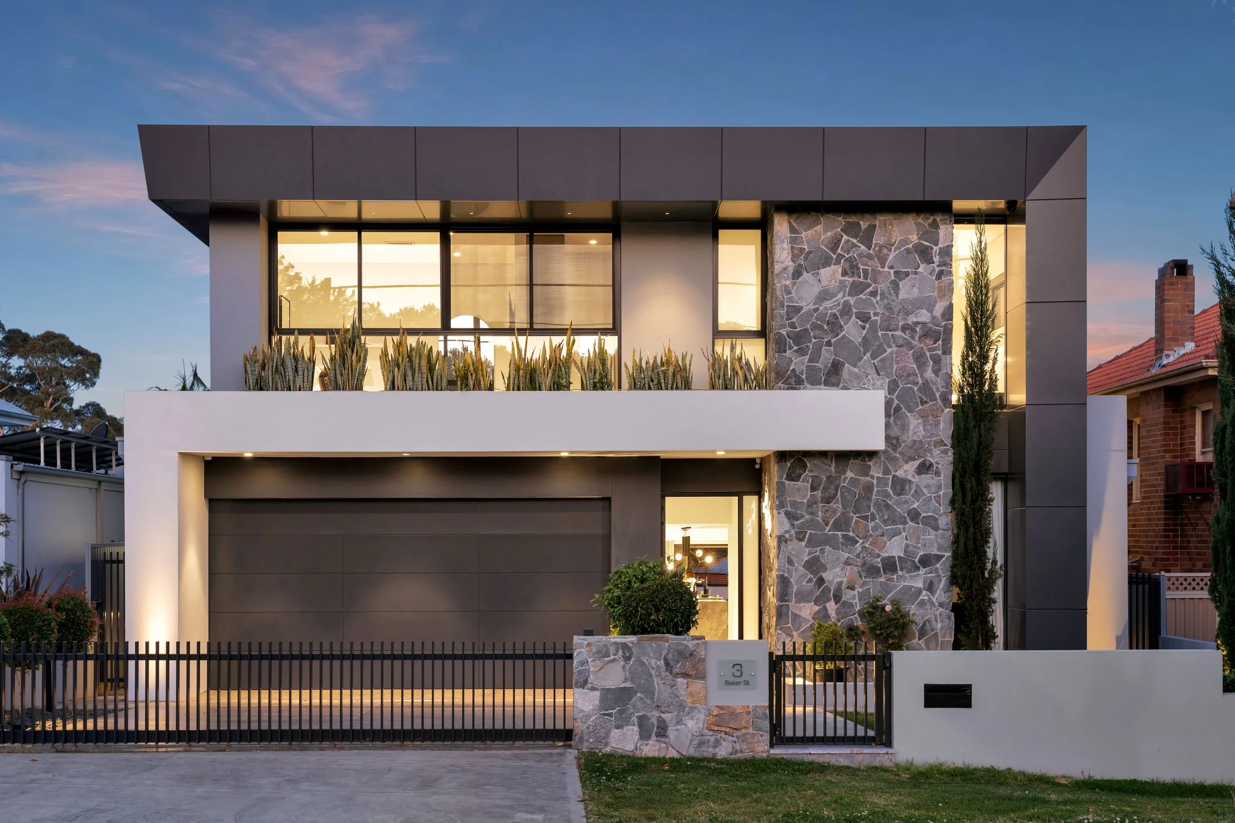 Modern two-story house with a stone and dark panel exterior, large windows, a garage, and a small front yard at dusk.