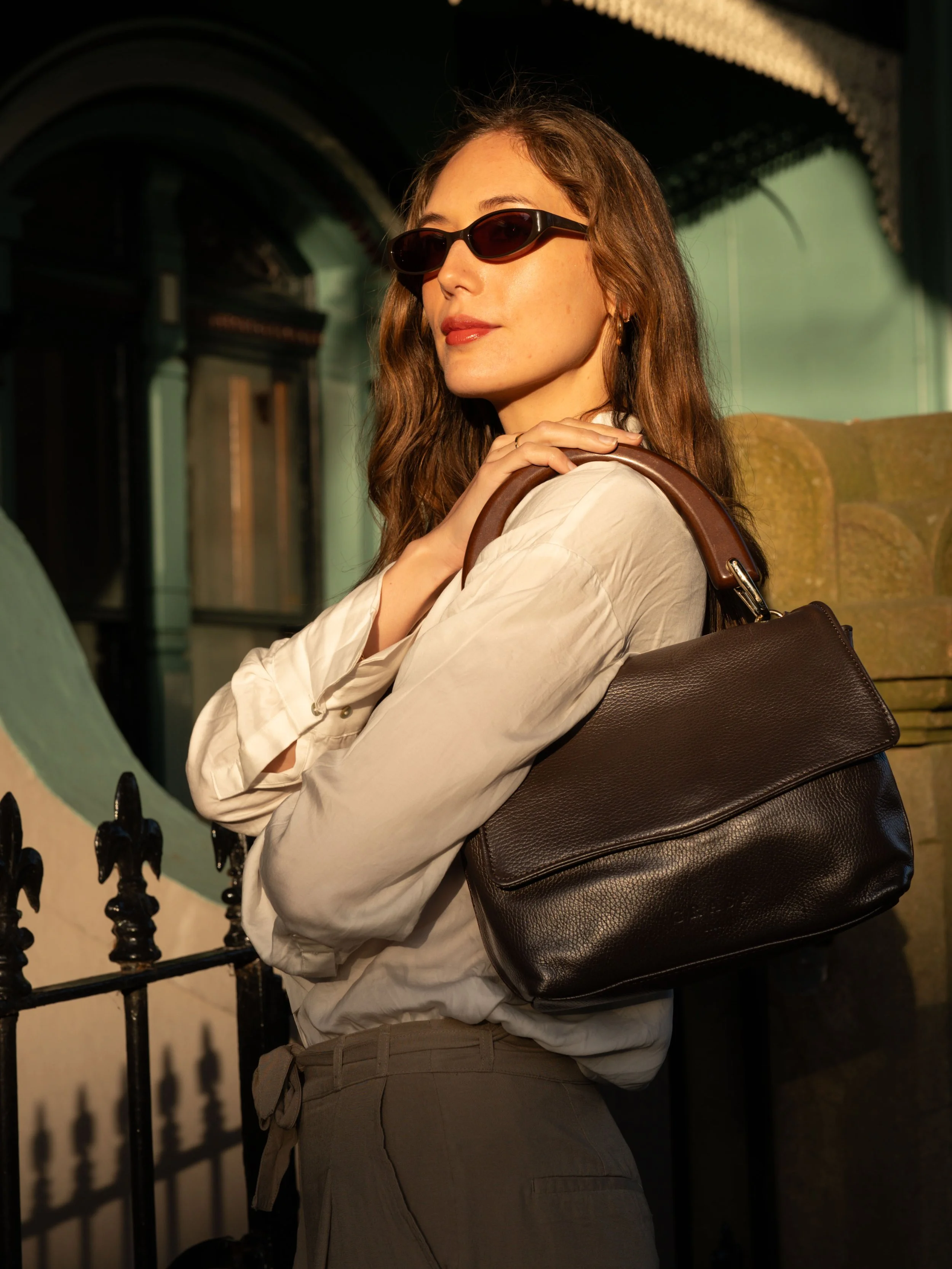 A woman with long brown hair wearing black sunglasses, a white shirt, and beige pants, carrying a black leather handbag on her shoulder, standing in front of a building with large windows and a black iron fence.