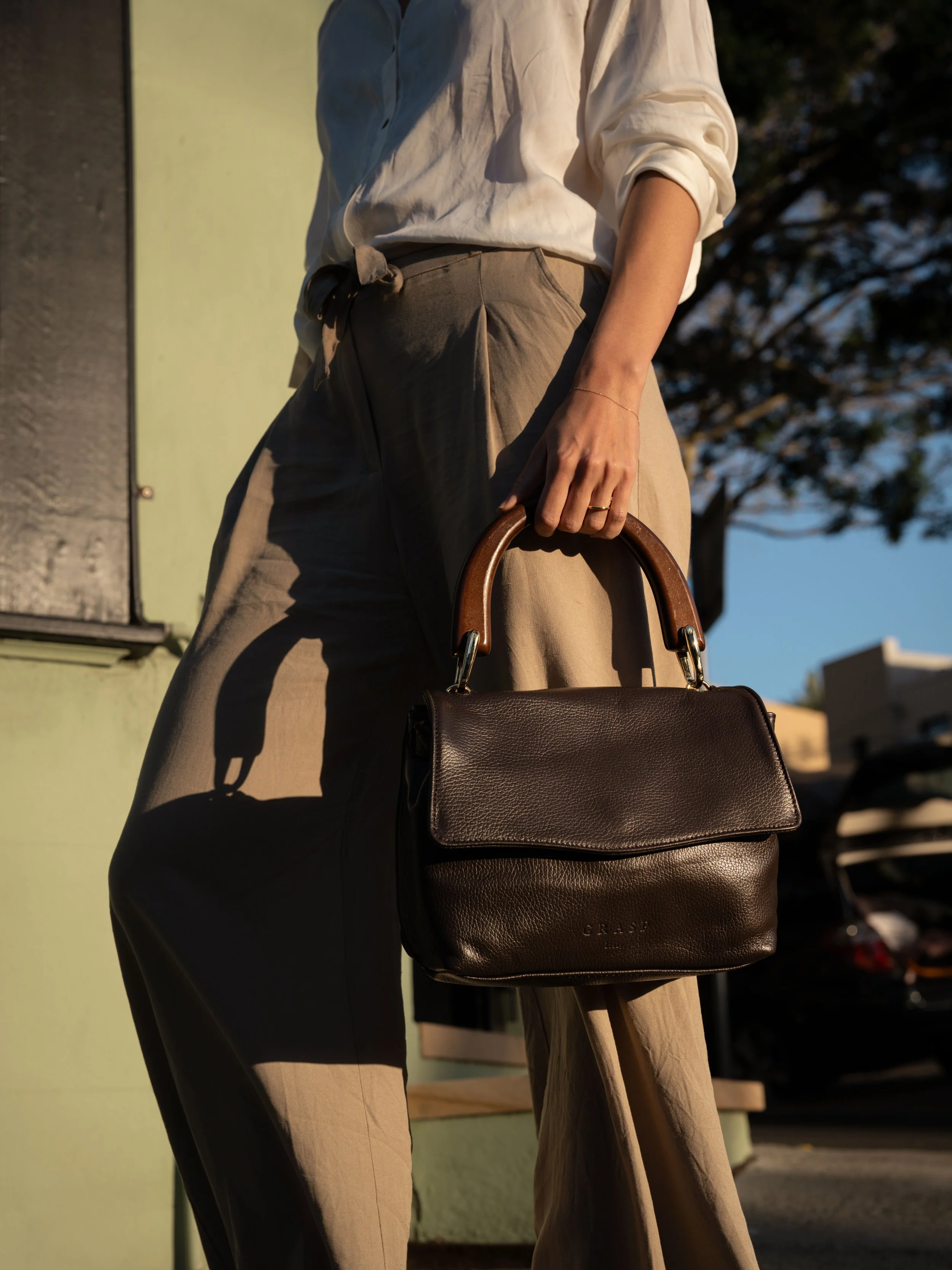 Person holding a black leather handbag with a tan handle, dressed in beige pants and a white shirt, standing outdoors during sunset.