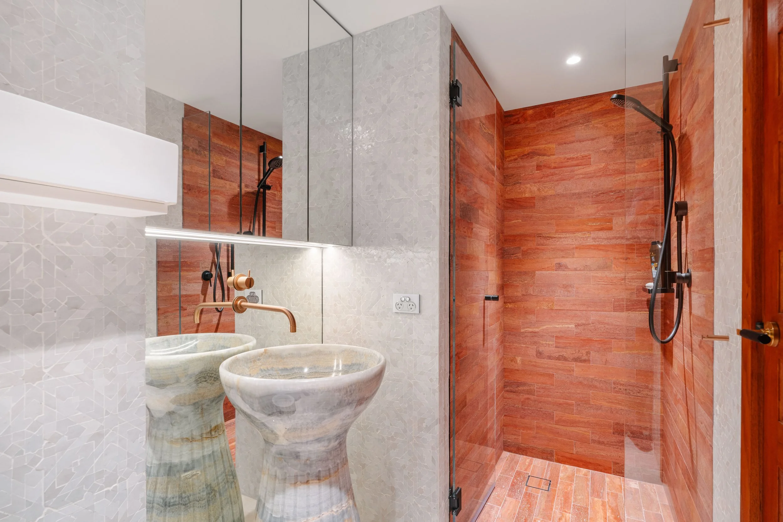 Modern bathroom with a shower area featuring wood-like tiles, a glass door, and pebble-style sinks with copper faucets.