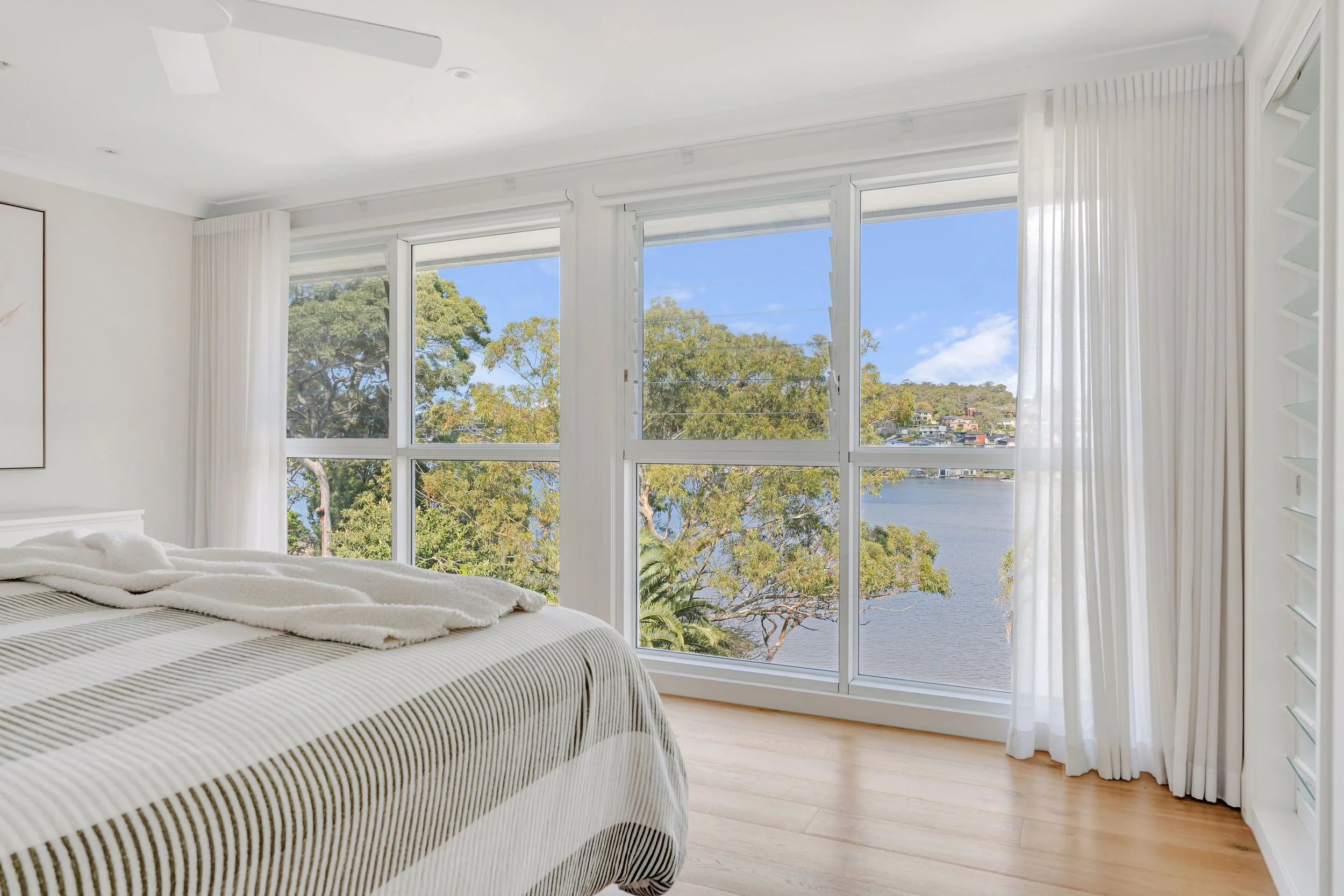 Bedroom with large window view of trees and a river, white curtains, light wood flooring, and a bed with a striped comforter.
