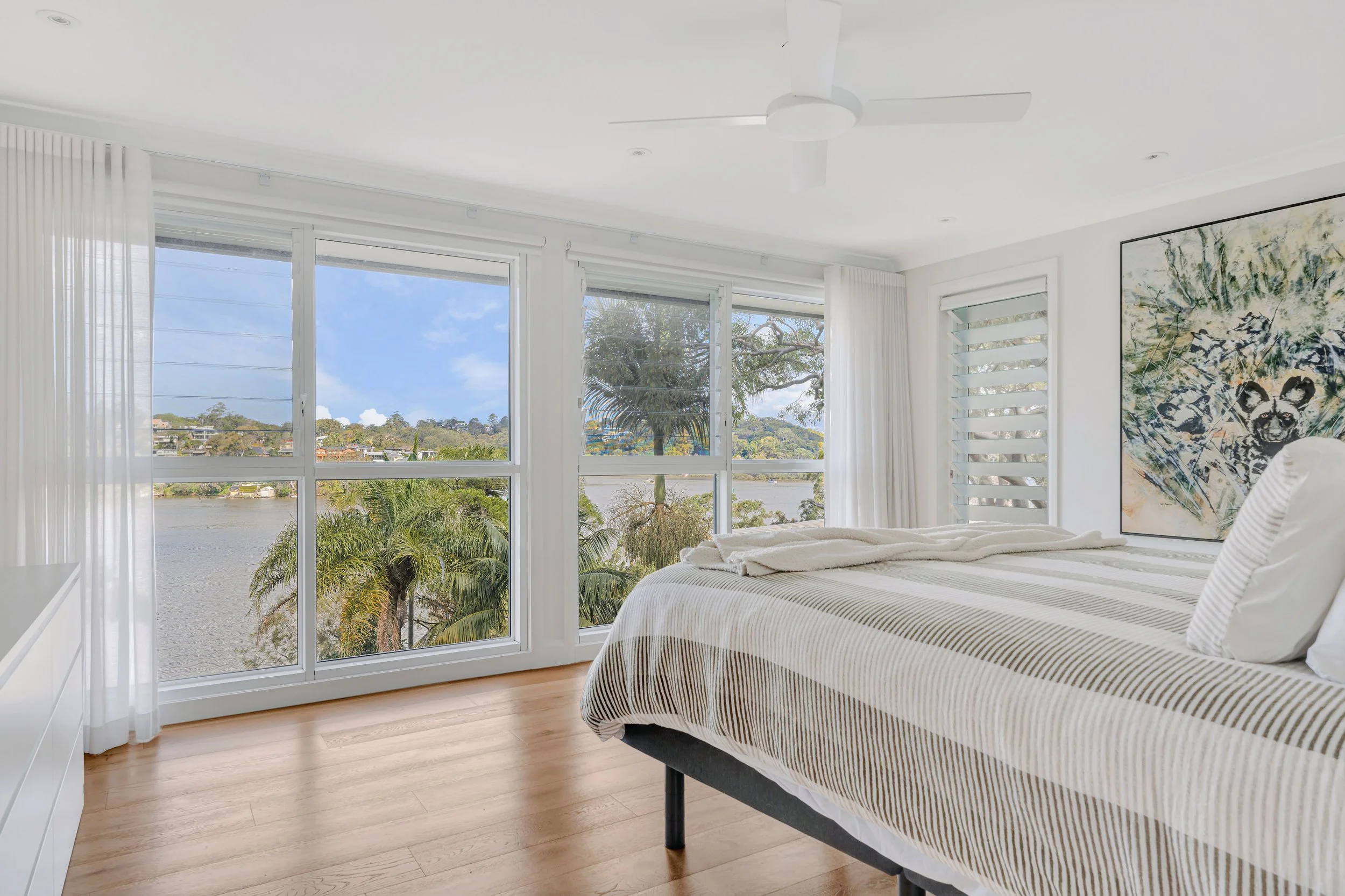 Bedroom with large windows showing a view of trees, water, and a partly cloudy sky, light-colored hardwood floor, white walls, white curtains, and a bed with a striped bedspread and pillows.