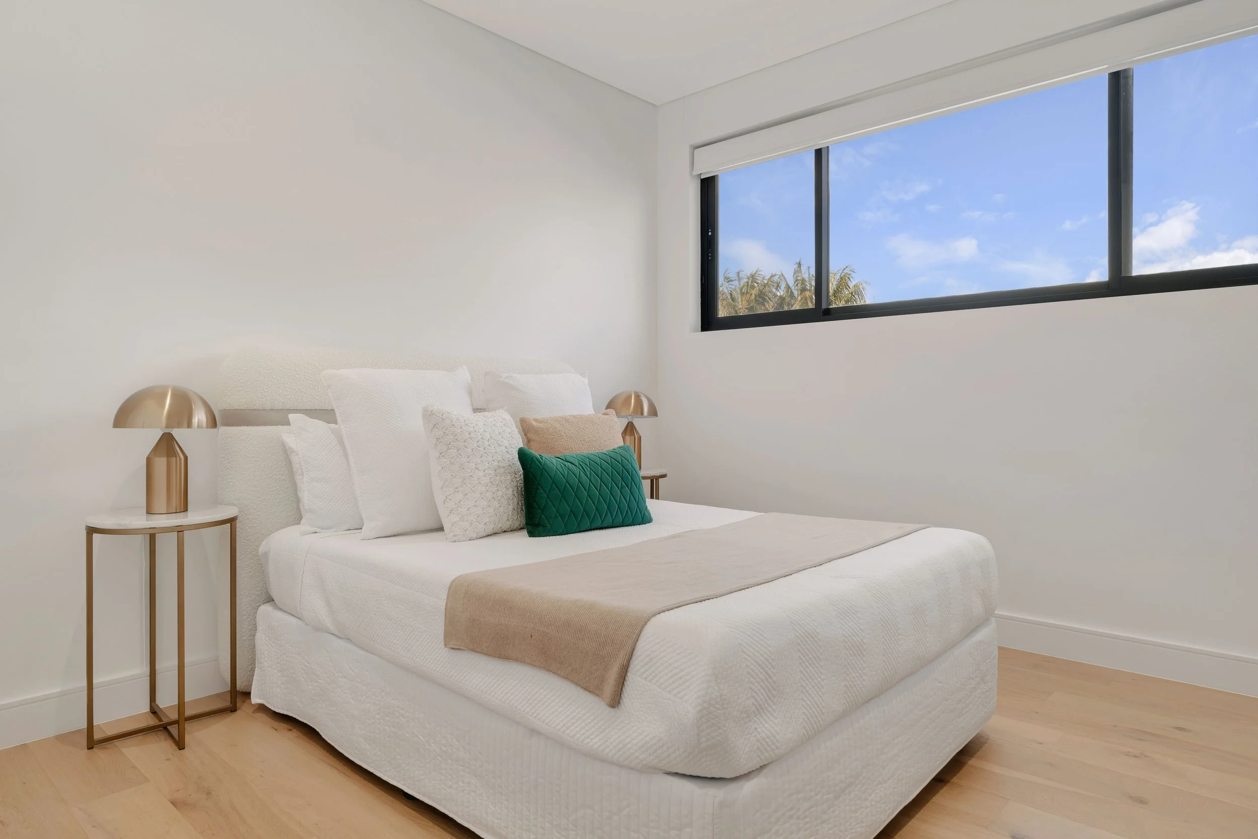 Minimalist bedroom with white bed, multiple pillows, beige throw blanket, two gold bedside lamps, and window with view of blue sky and trees.